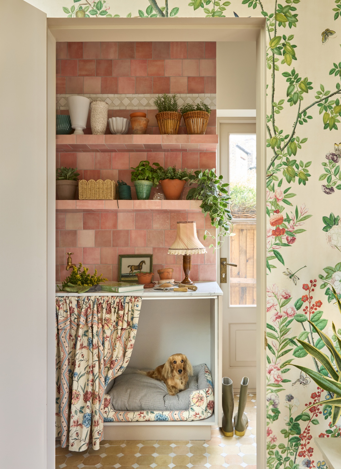 A wall with cream and green botanical wallpaper looking into a boot room with a dog bed alcove with a dachsund laying on a bed cover with a floral curtain