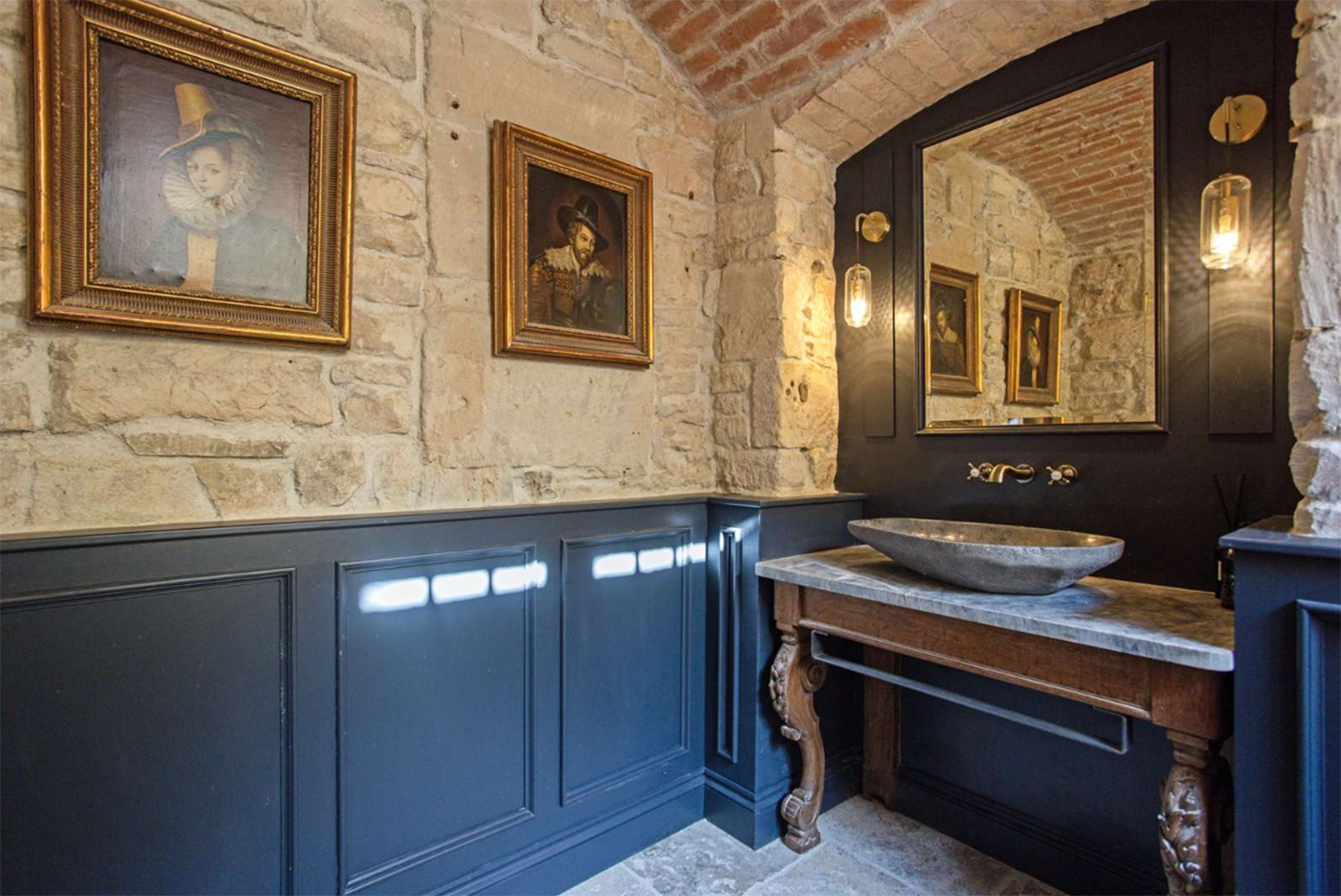 a bathroom with exposed stone walls, blue painted panelled walls, a marble basin with wooden legs and portrait paintings in gilded frames