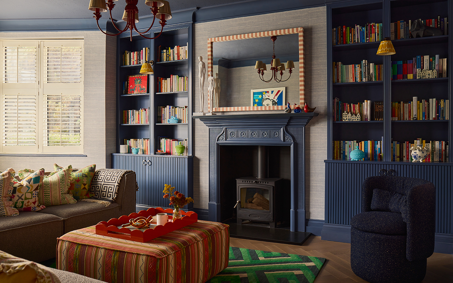 a living room with a fireplace and built in bookcases in a painted blue finish. A corner sofa and accent chair sit around a patterned ottoman and graphic rug. A stripy mirror hangs on the wall. A mulit-arm chandelier hangs from the ceiling