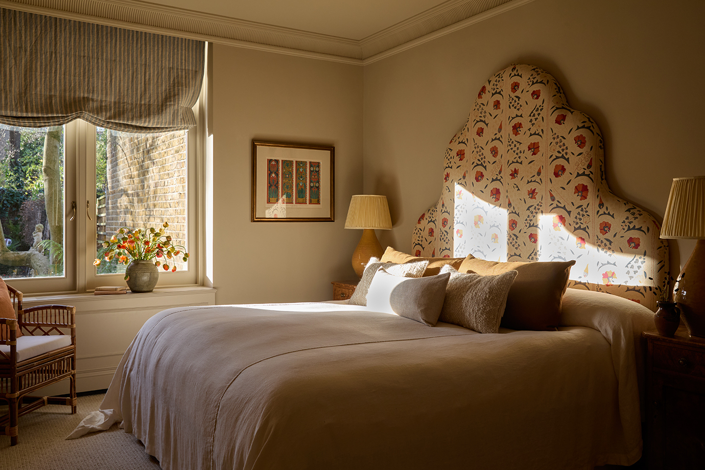 A bedroom with a double bed finished with a large statement headboard in a floral fabric. On the left a window is dressed with a striped relaxed blind