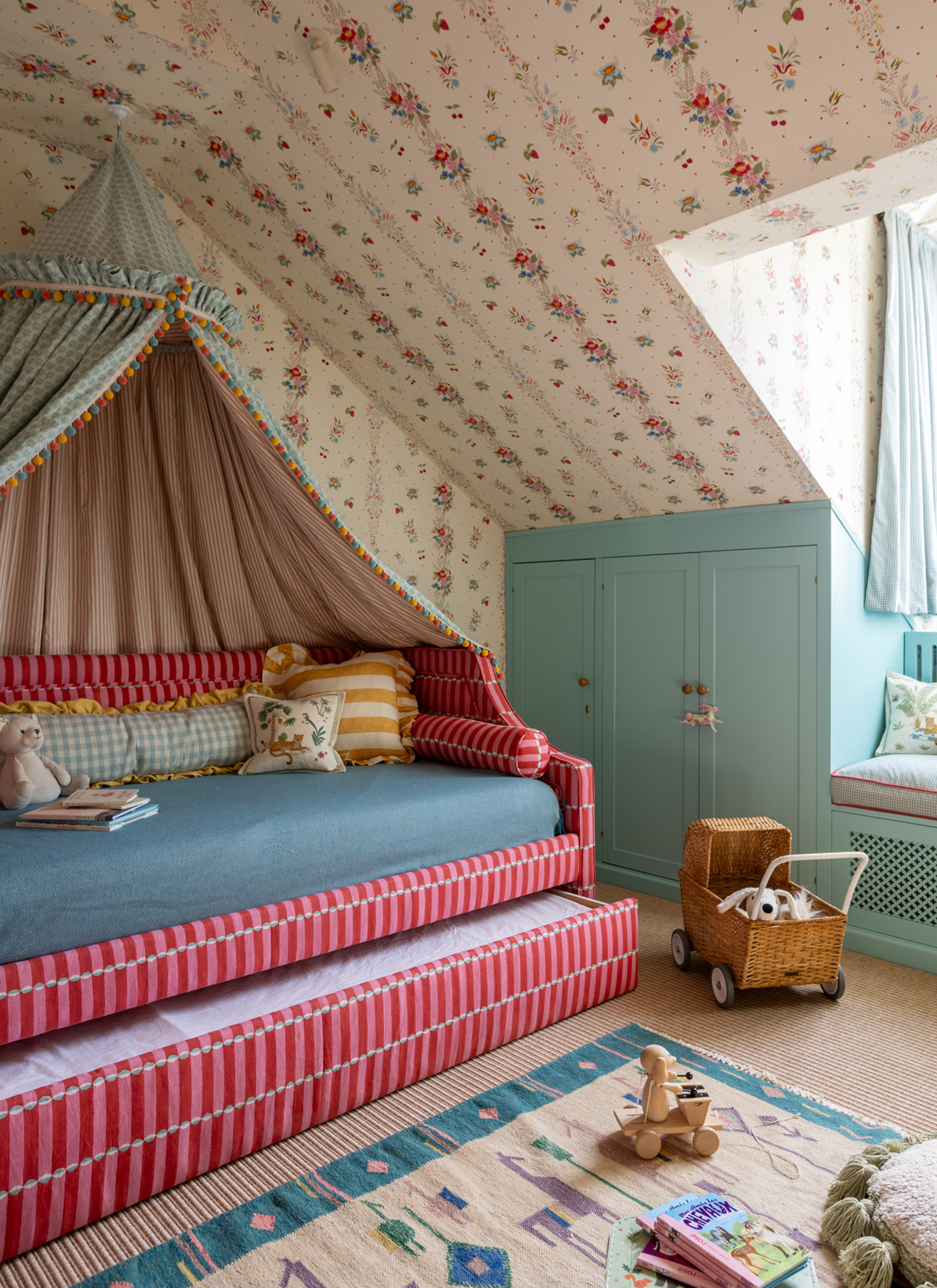 a children's bedroom with floral wallpaper and a large red and pink stripe daybed with a pull out storage drawer and a blue canopy with pom pom trim