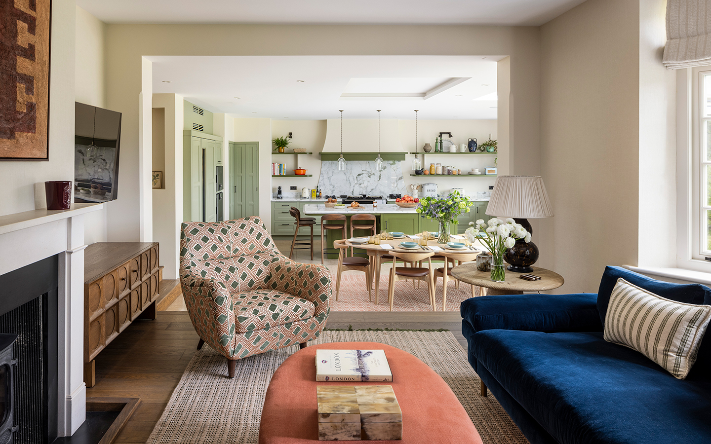 an open-plan area in a family home with a living area, with an armchair, sofa and ottoman, in the foreground, a wooden dining table and set of chairs in the middle and a green and marble kitchen in the background