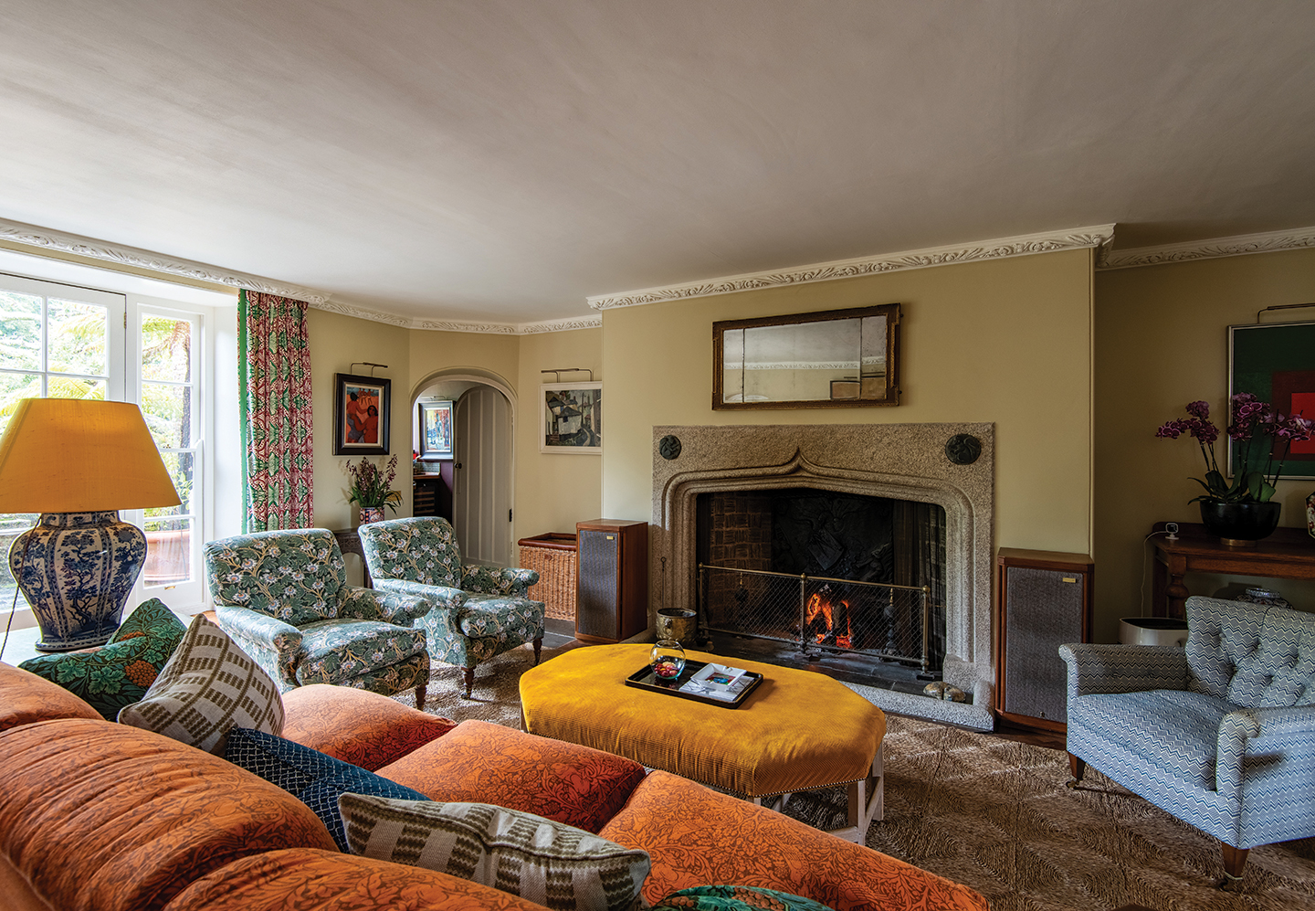 a traditional living room with orange sofa and three blue patterned armchairs surrounding a mustard yellow corduroy ottoman. A jute rug covers the floor and a fire is lit in a stone fireplace