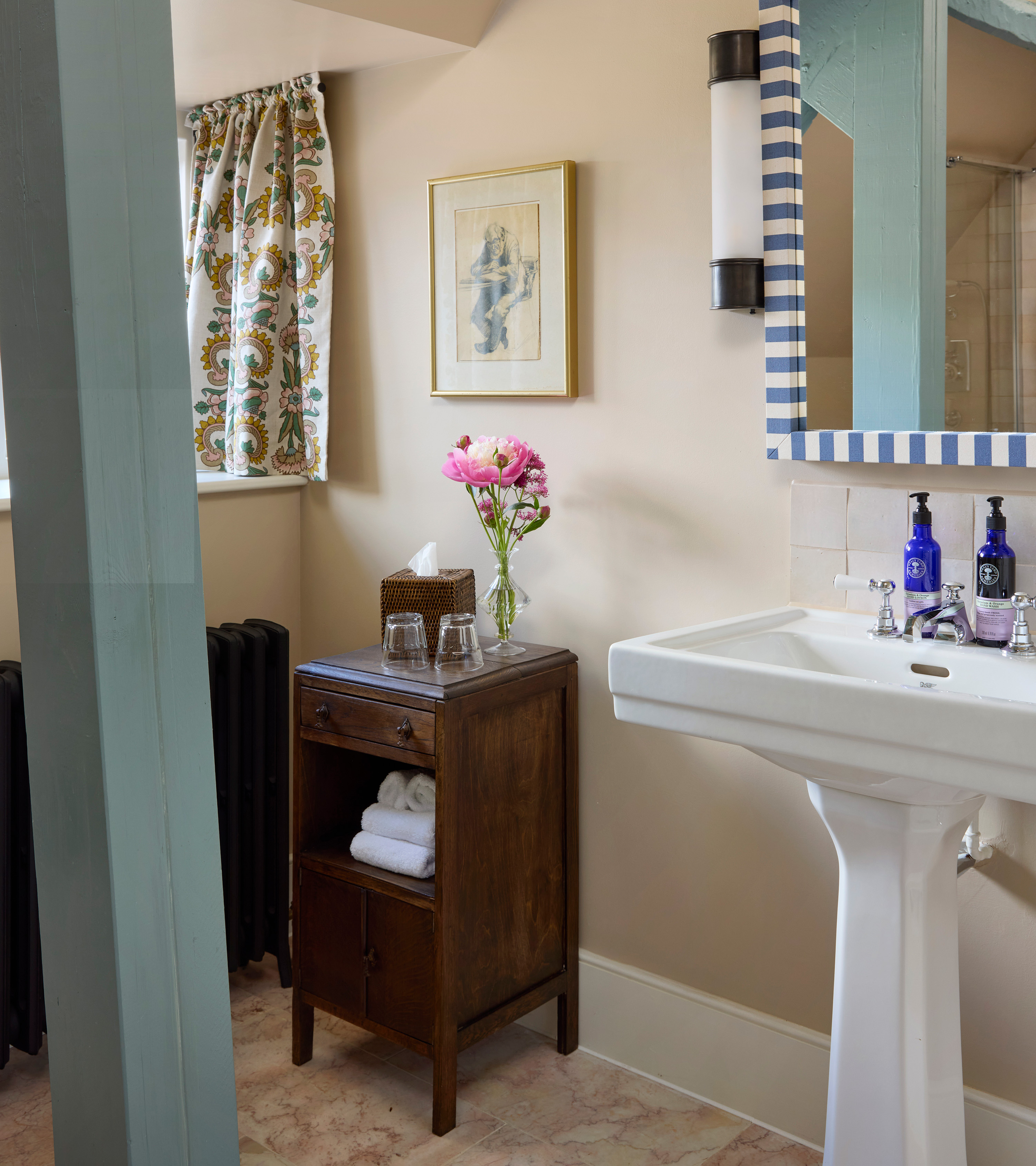 a bathroom corner with an antique dark wood storage unit with a vase and pink flowers on top, a white sink with a blue and white striped mirror above