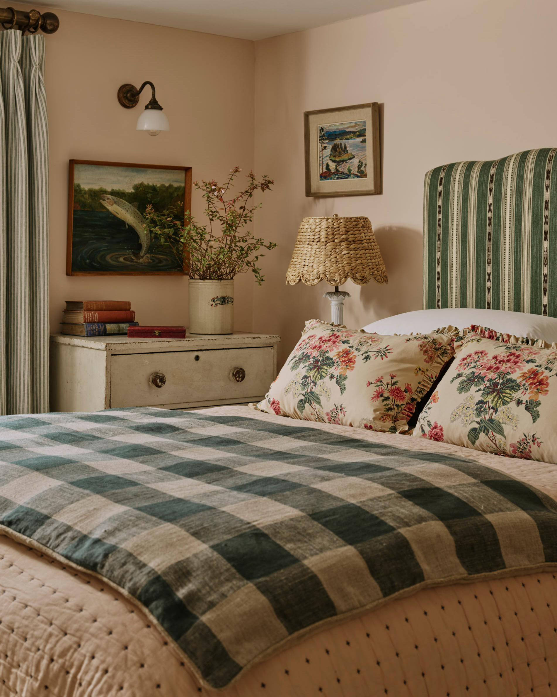 a bedroom with plaster pink walls, a bed with a green and cream stripe headboard and a green and cream gingham bedspread, a rattan lampshade and a white painted chest of drawers next to the bed