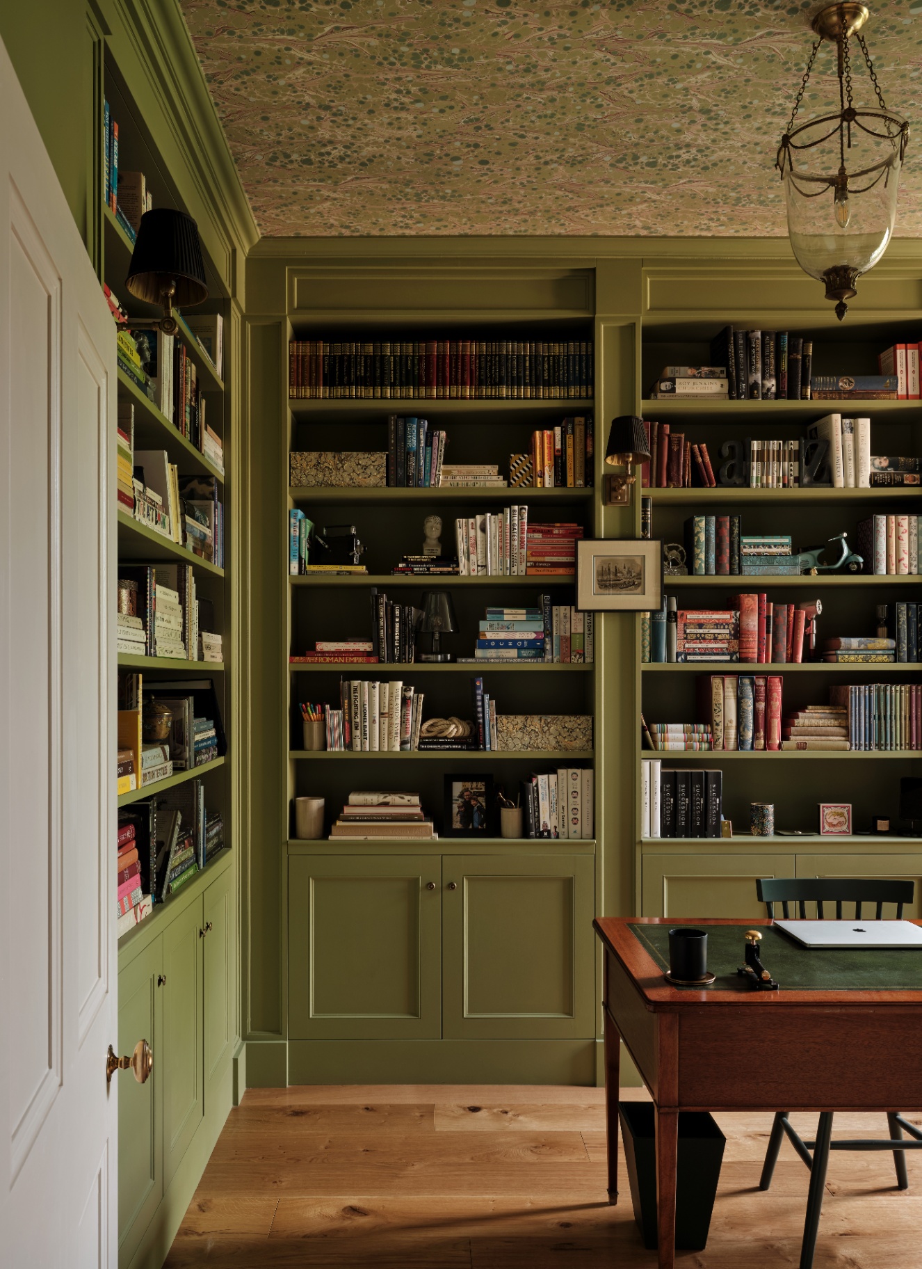 Olive green built-in library cabinetry surrounding a workspace, featuring a marbled patterned wallpaper applied across the ceiling and a brass lantern light fixture