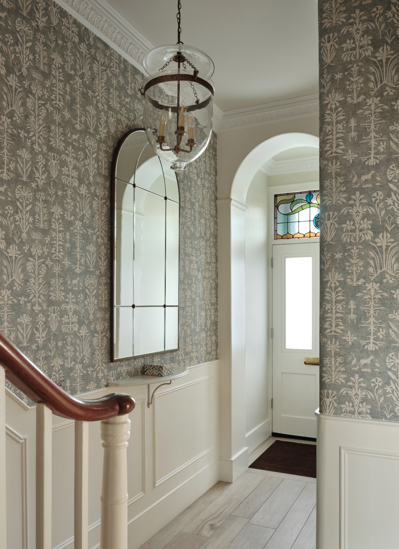 a hallway with sage and white botanical wallpaper, white panelling, an arched mirror and a large lantern pendant light