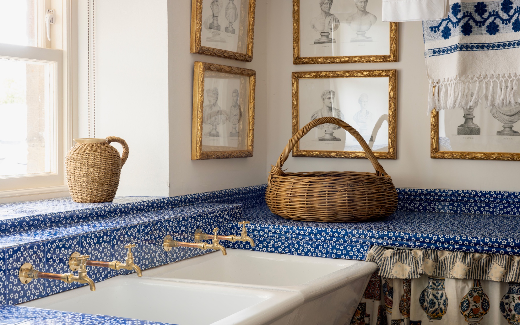 a utility room sink with blue and pink floral tiles, a set of pencil sketches framed on the walls and a wicker basket on the countertop