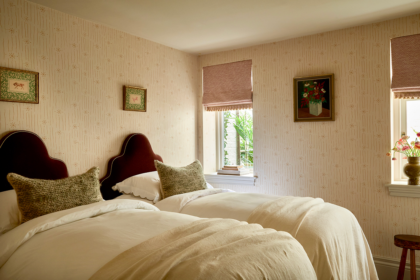 a twin bedroom wallpapered in a delicate yellow design. The two single beds are dressed with neutral linens and textured pillows and have shapely headboards covered in a reddish-brown velvet. Two small windows are dressed with tasselled pink blinds