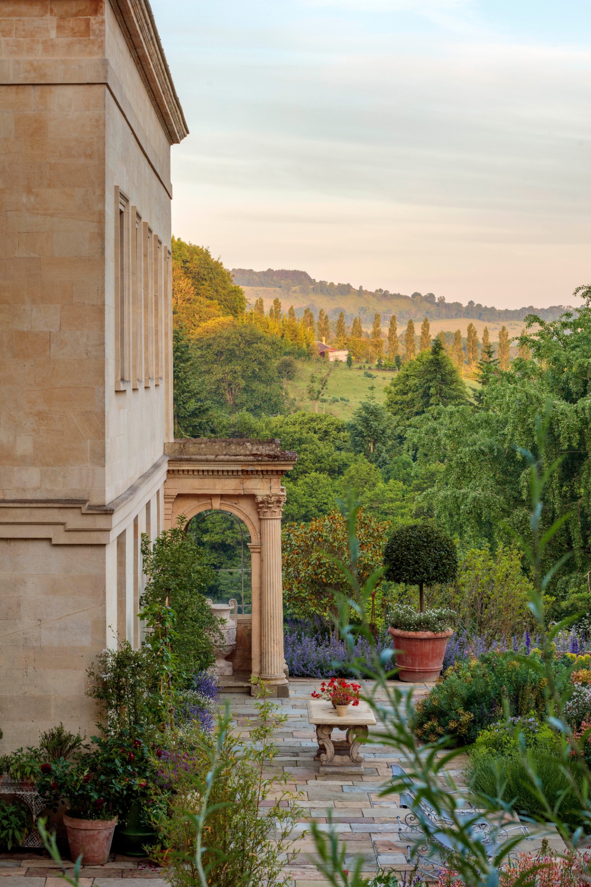 Exterior of Jade Holland Cooper’s neoclassical Bath stone estate with a stone patio, potted plants, and sweeping Cotswolds countryside views