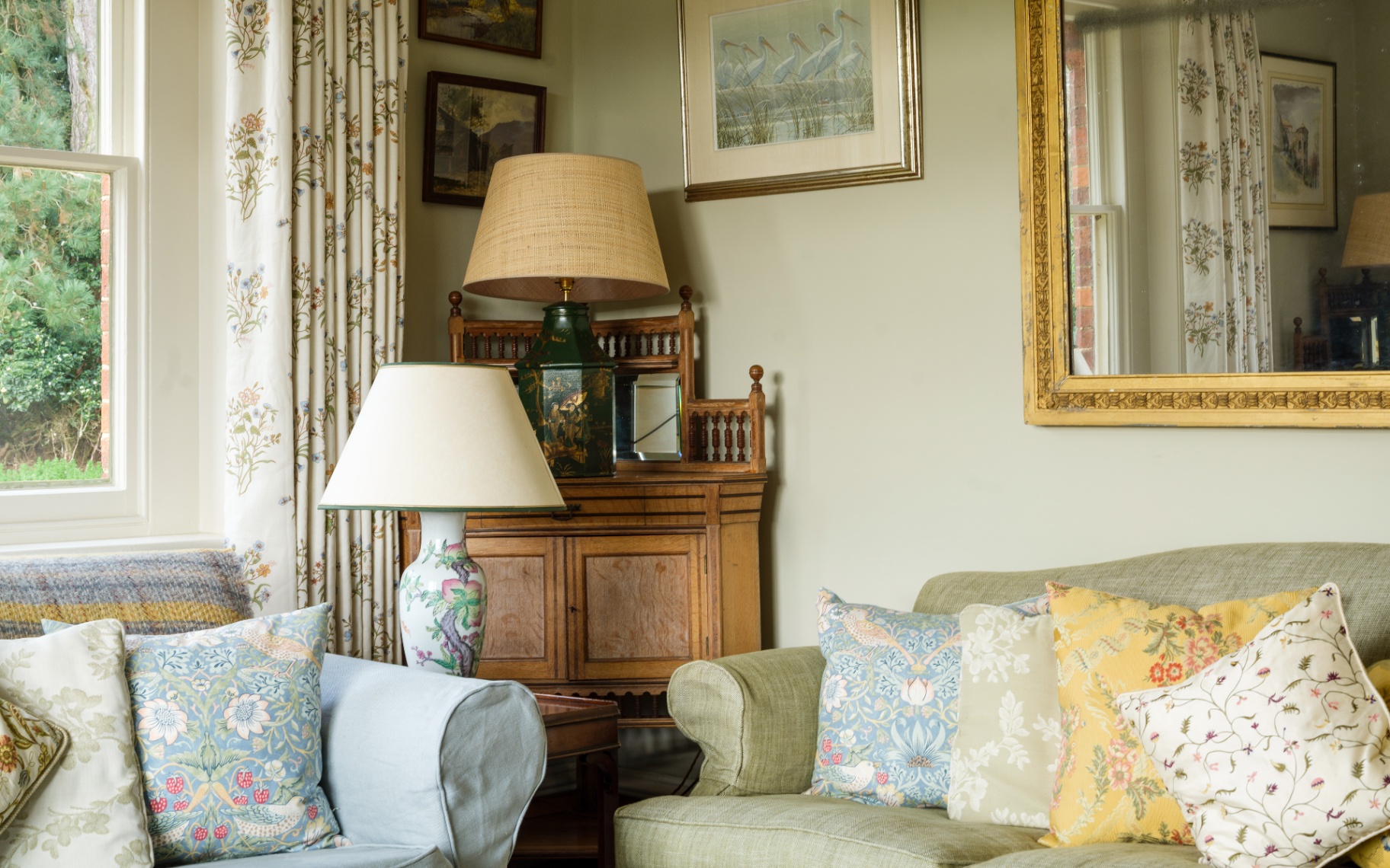 a country theme sitting room with pale green walls, a large gold ornate mirror and framed art on the walls and two sofas with lots of floral cushions