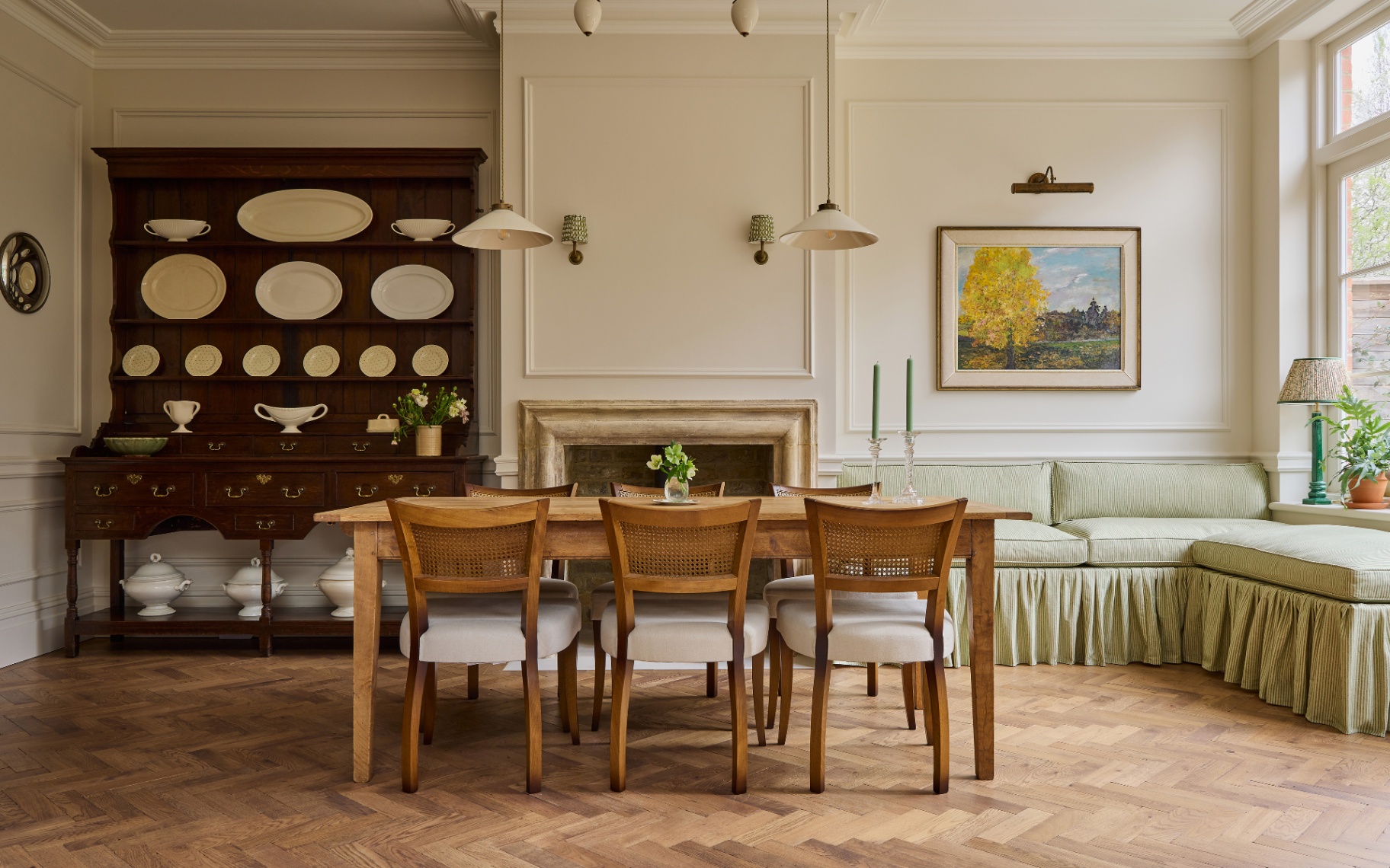 a neutral dining room with a wooden dining table, a green skirted sofa to the right with a large piece of art hanging above it and a large dark wooden dresser filled with decorative plates to the left