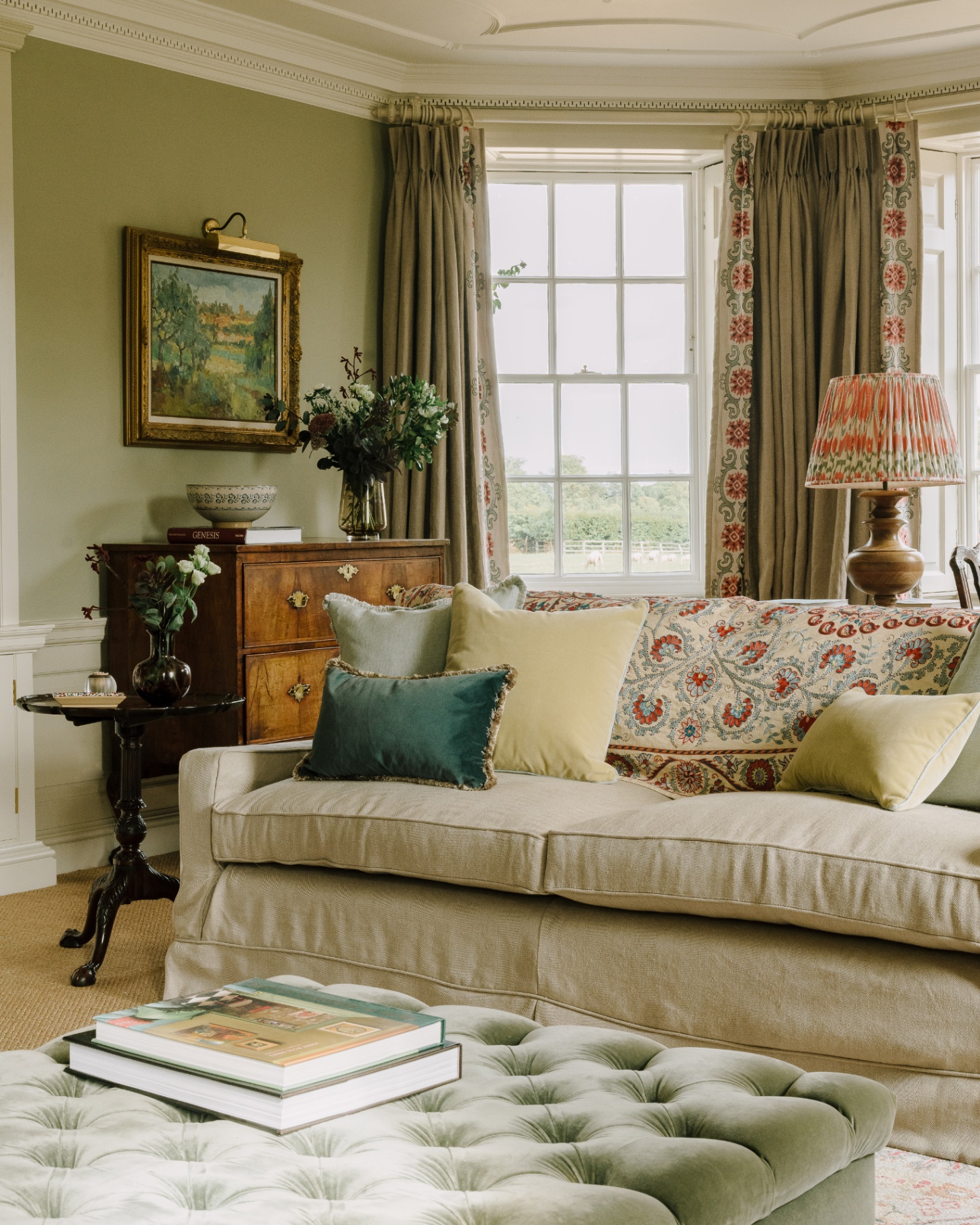 a living room with a cream linen sofa with blue and pale yellow cushions, a sage green velvet ottoman and long beige curtains behind the sofa