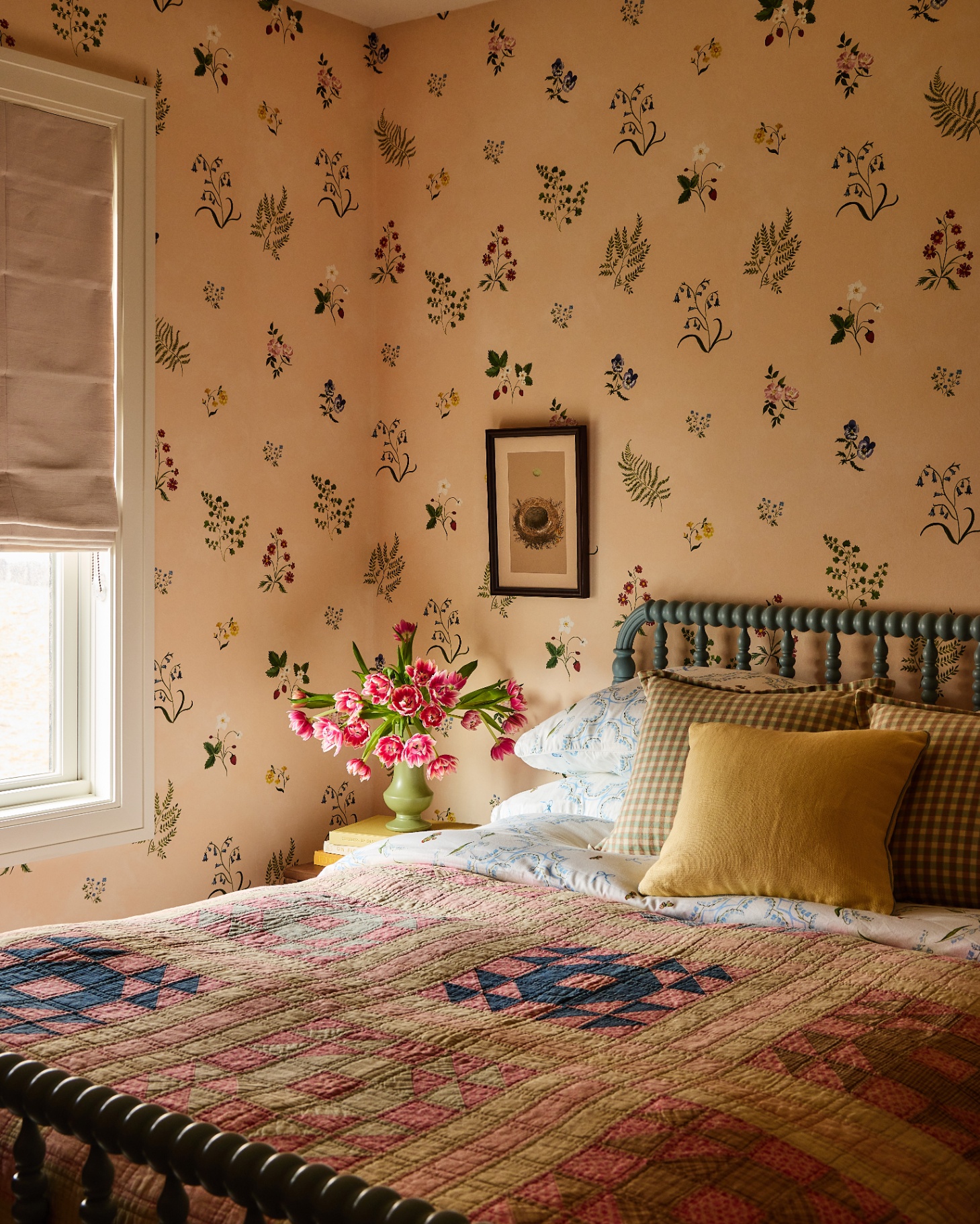 a bedroom with ochre walls with small painted flowers, an antique bobbin bed with a yellow, blue and pink patchwork quilt and a vase of pink flowers on a bedside table