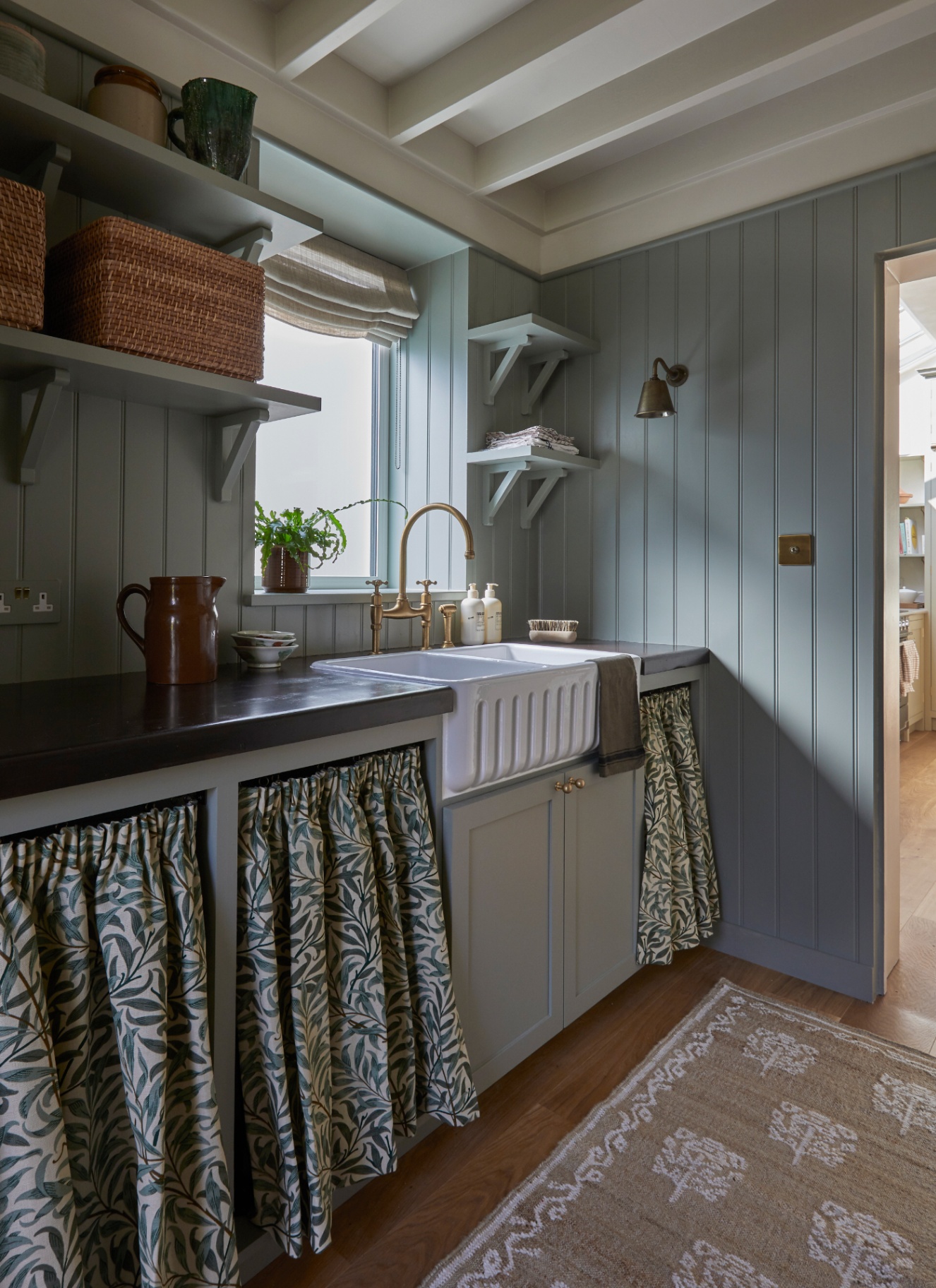 a utlity room with pale blue walls, open shelves with towels and baskets, a belfast sink and a white and blue leaf patterned sink skirt