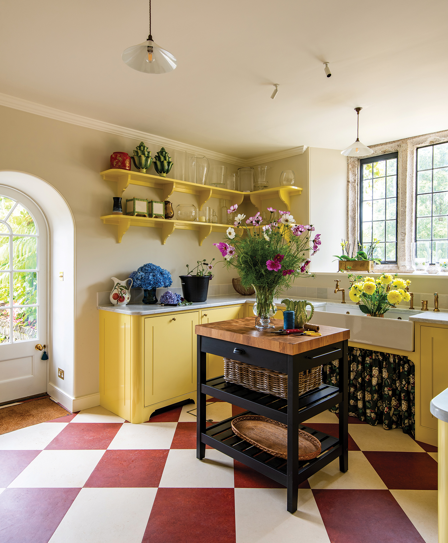 A flower room with red and white checkerboard floor and bright yellow cabinets and shelving. In the centre is a mini island with black-painted legs and shelves and a wooden top. A butler sink has a vase of flowers in it and a floral sink skirt beneath