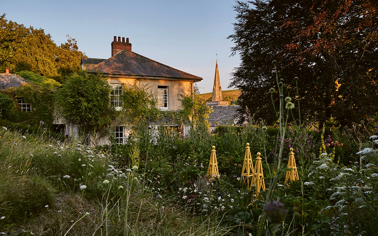 A 19th-century building covered in climbing plants, with a cottage-style garden in front. In the background are a church spire, fields and trees