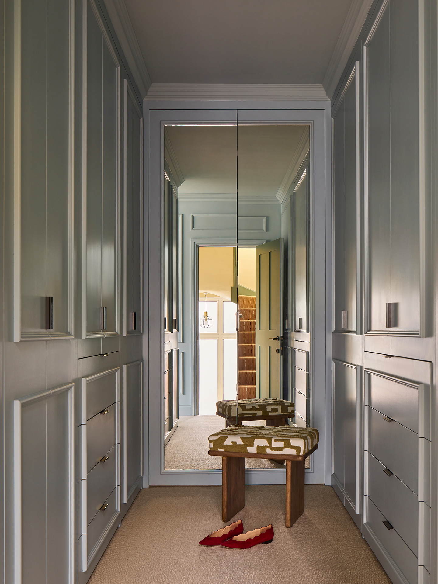 a light blue narrow dressing room with built-in joinery either side and a floor to ceiling mirror at the end. In front of the mirror sits a wooden stool with an upholstered seat in a graphic fabric and a pair of flat shoes 