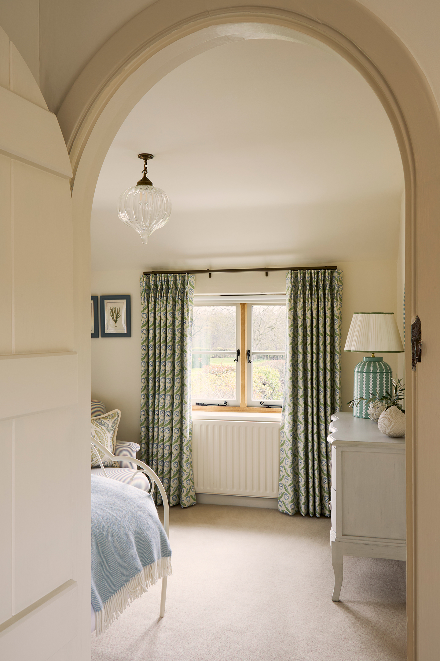 A view through an arched doorway into a neutral bedroom with the corner of a bed visible and a chest of drawers topped with a lamp and vase. On the far wall a radiator sits below a window dressed with floral blue and green curtains 