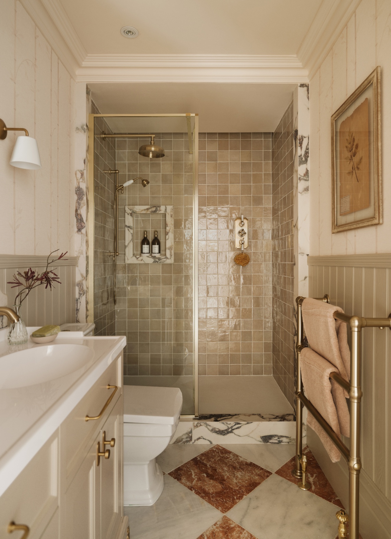a bathroom with awalk in shower with beige tiles and gold hardware, a white and rust checkerboard marble floor, half white and half beige paneled walls and a cream double sink vanity