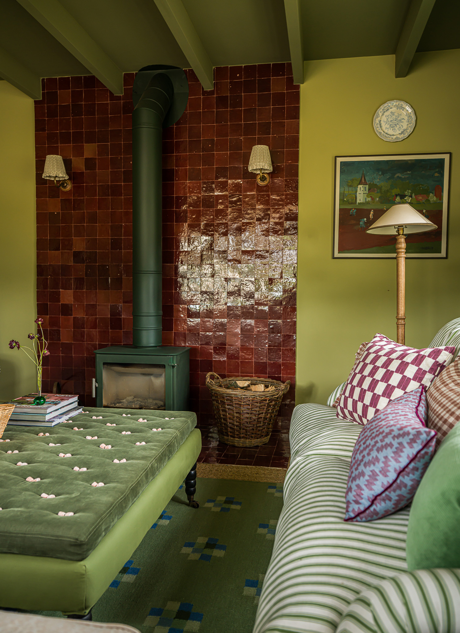 A cozy olive-green living area with a green log burner against glossy terracotta-tiled walls, a green velvet tufted ottoman, and a striped sofa with patterned cushions.