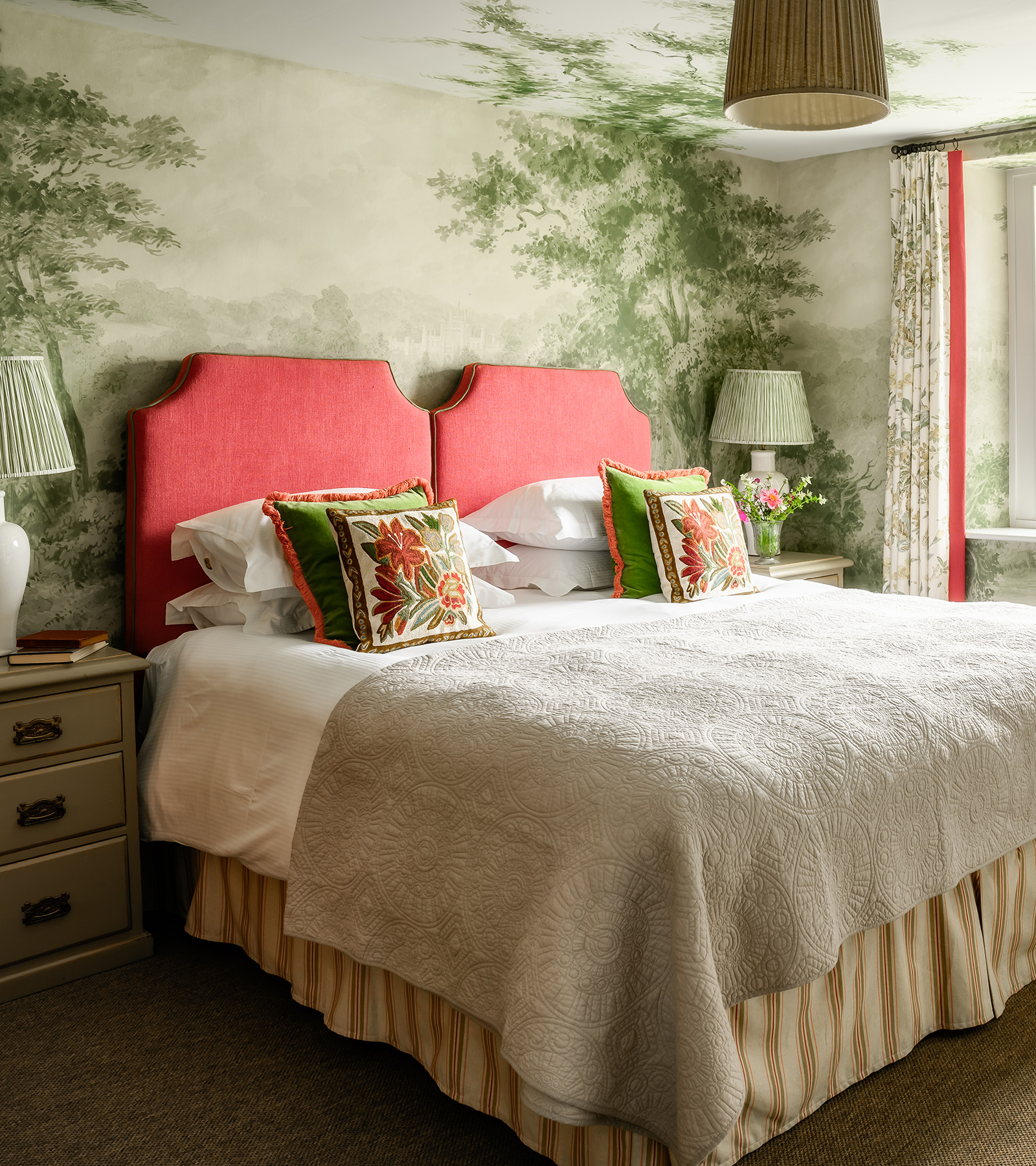 a bedroom with green and white mural wallpaper featuring trees with a double bed with a red headboard 