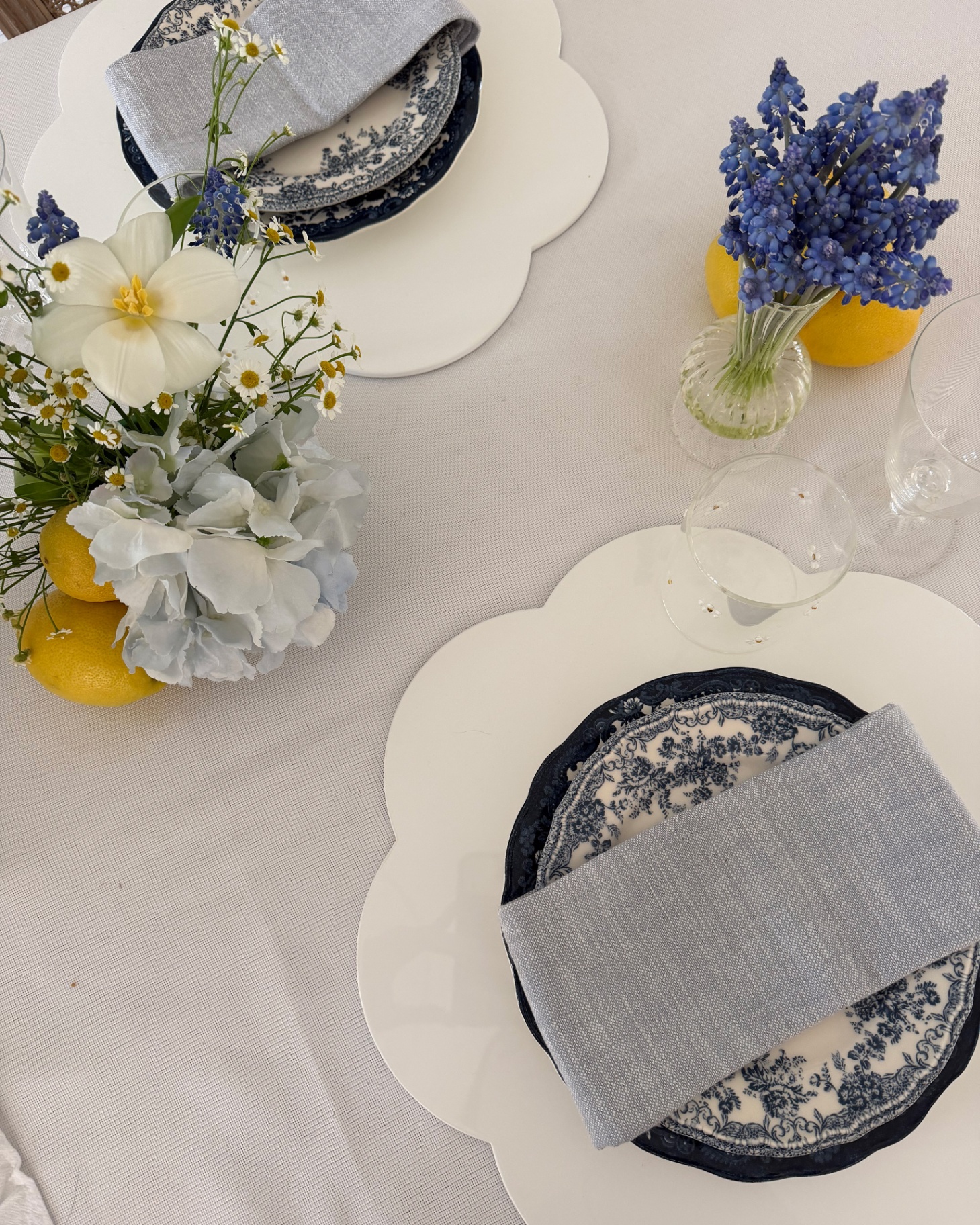 a white spring tablescape with a white tablecloth, white placemats, clue and white patterned plates, blue napkins and bud vases filled with blue and yellow flowers