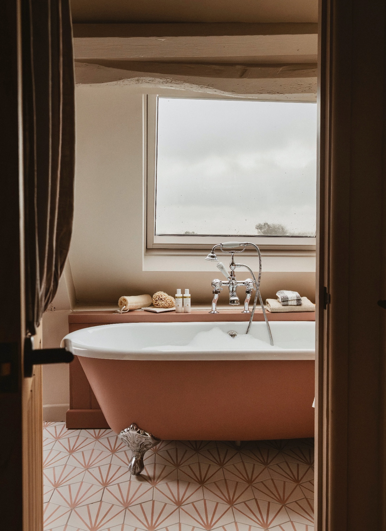 a bathroom with a salmon pink freestanding bath, a white and salmon pink patterned tiled floor and a large window