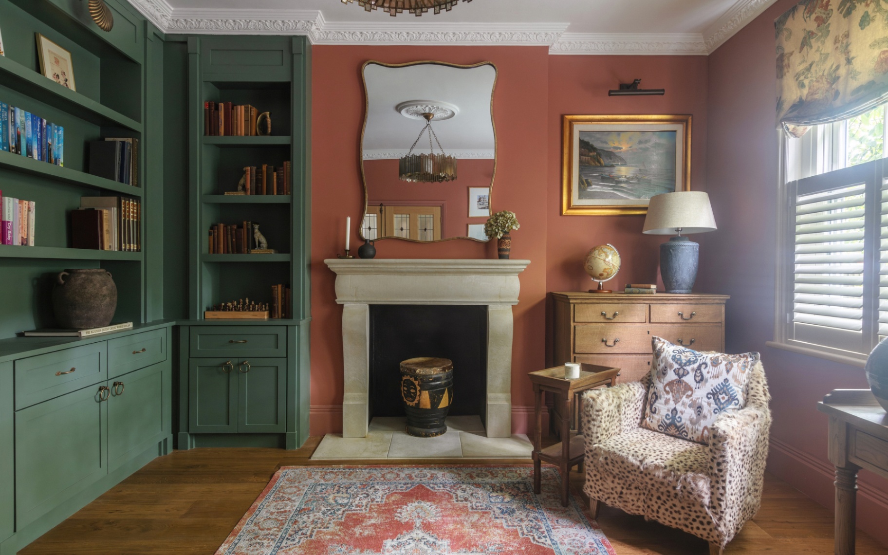a living room with rust orange walls, a small fluffy animal print accent chair, a built in corner bookcase painted in dark green and a a large stone fireplace with a wavy edge mirror above it