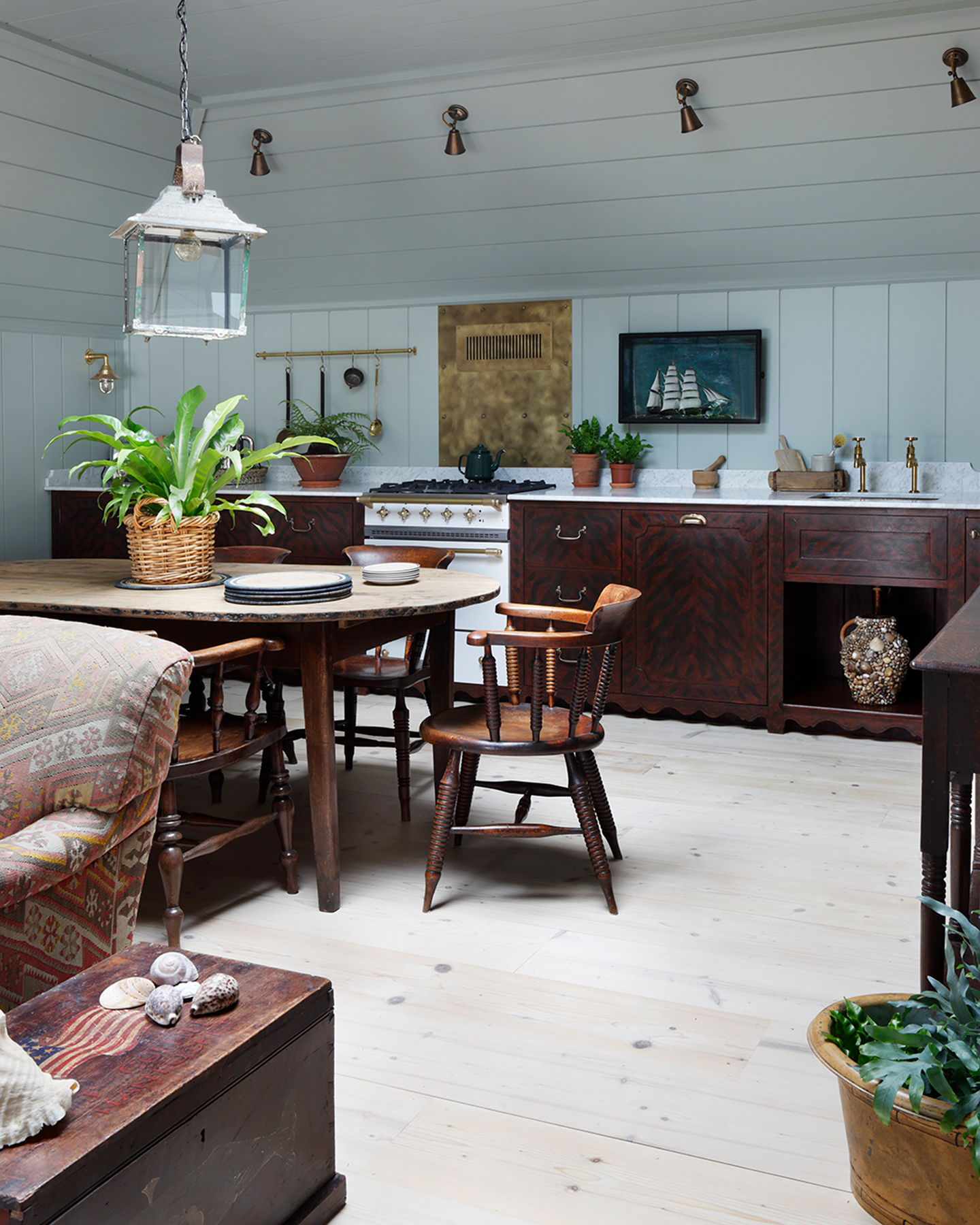 a kitchen with white floorboards, white panelled walls, 4 brass spotlights, a white and clear glass lantern pendant over a long oval wooden dining table and chairs and dark wood kitchen cabinets