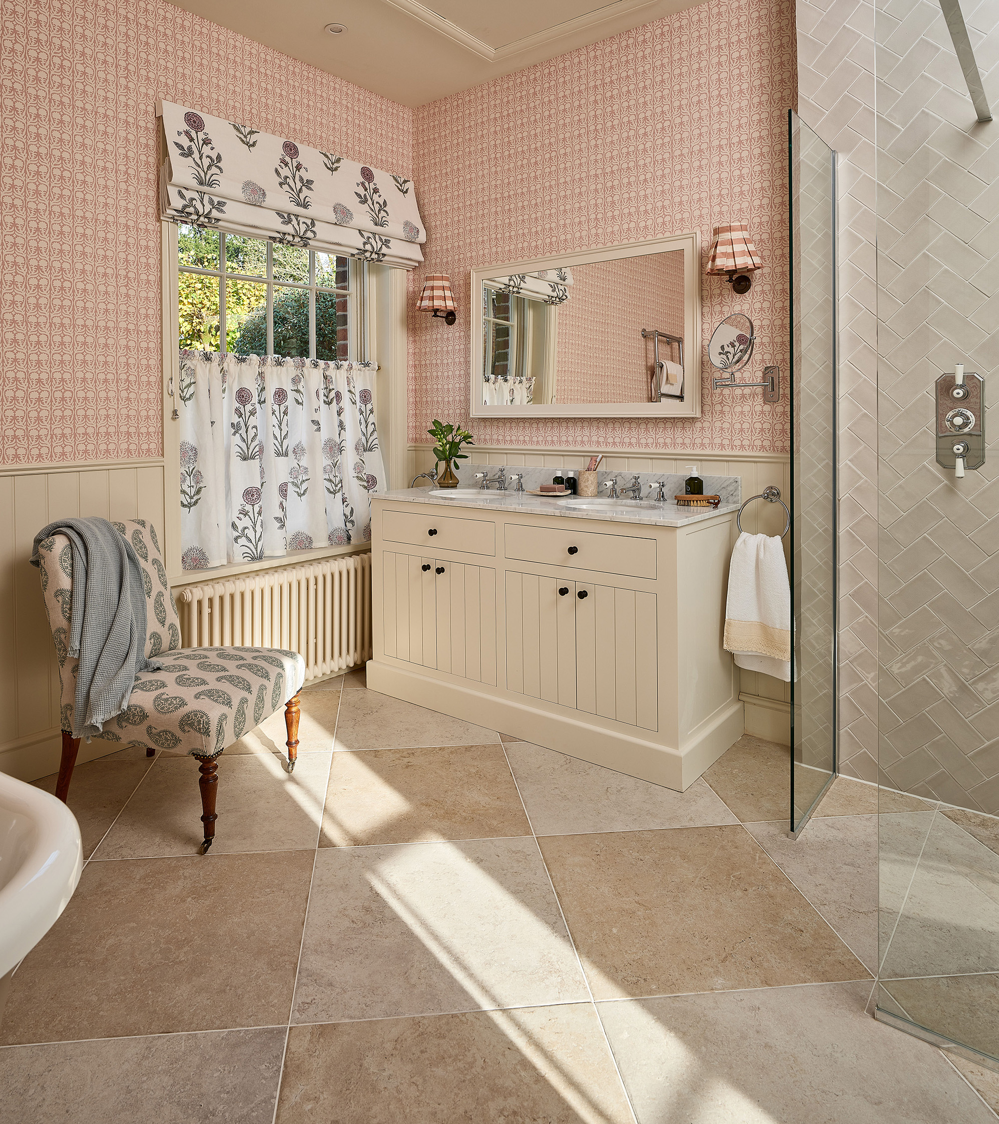 a bathroom with limestone floor, half cream panelled and half pink and white botanical wallpaper walls, a cream double vanity and sink with a ractangle mirror above and sconces either side