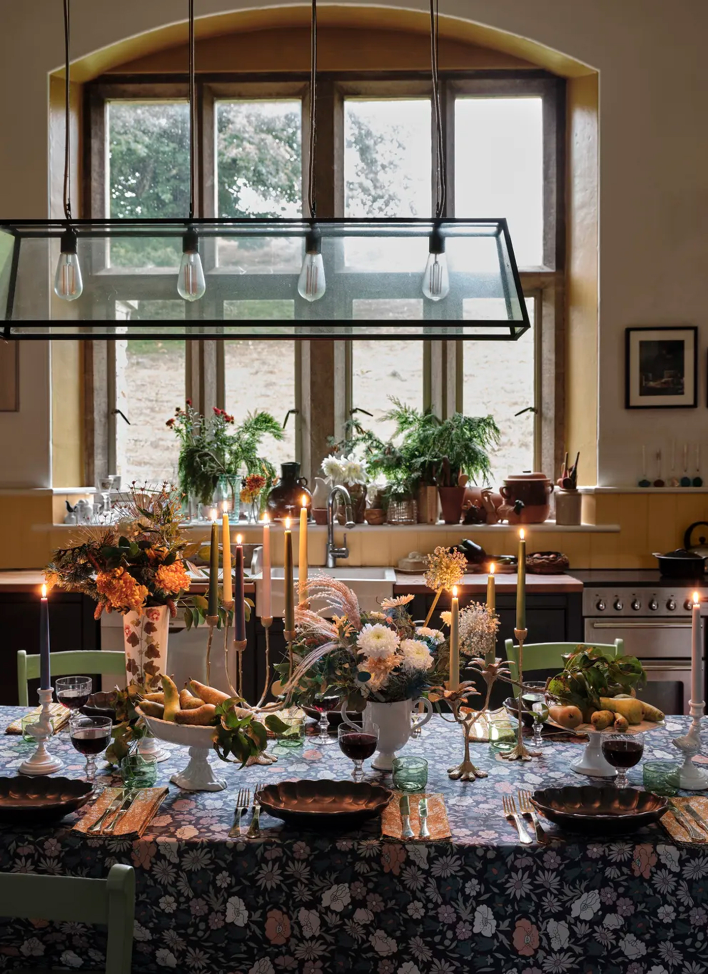 An autumn table in a kitchen with a large pendant lantern above it, lots of lit tapered candles, a dark floral tablecloth and vases of flowers
