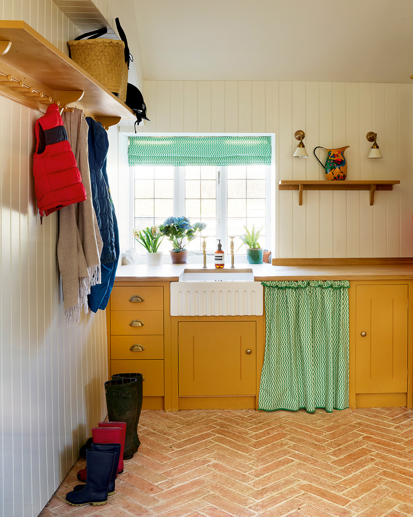 a utility room with tiled parquet floors, mustard yellow cabinetry, a green and white fabric skirt and matching blinds and a row of wellington boots on the floor
