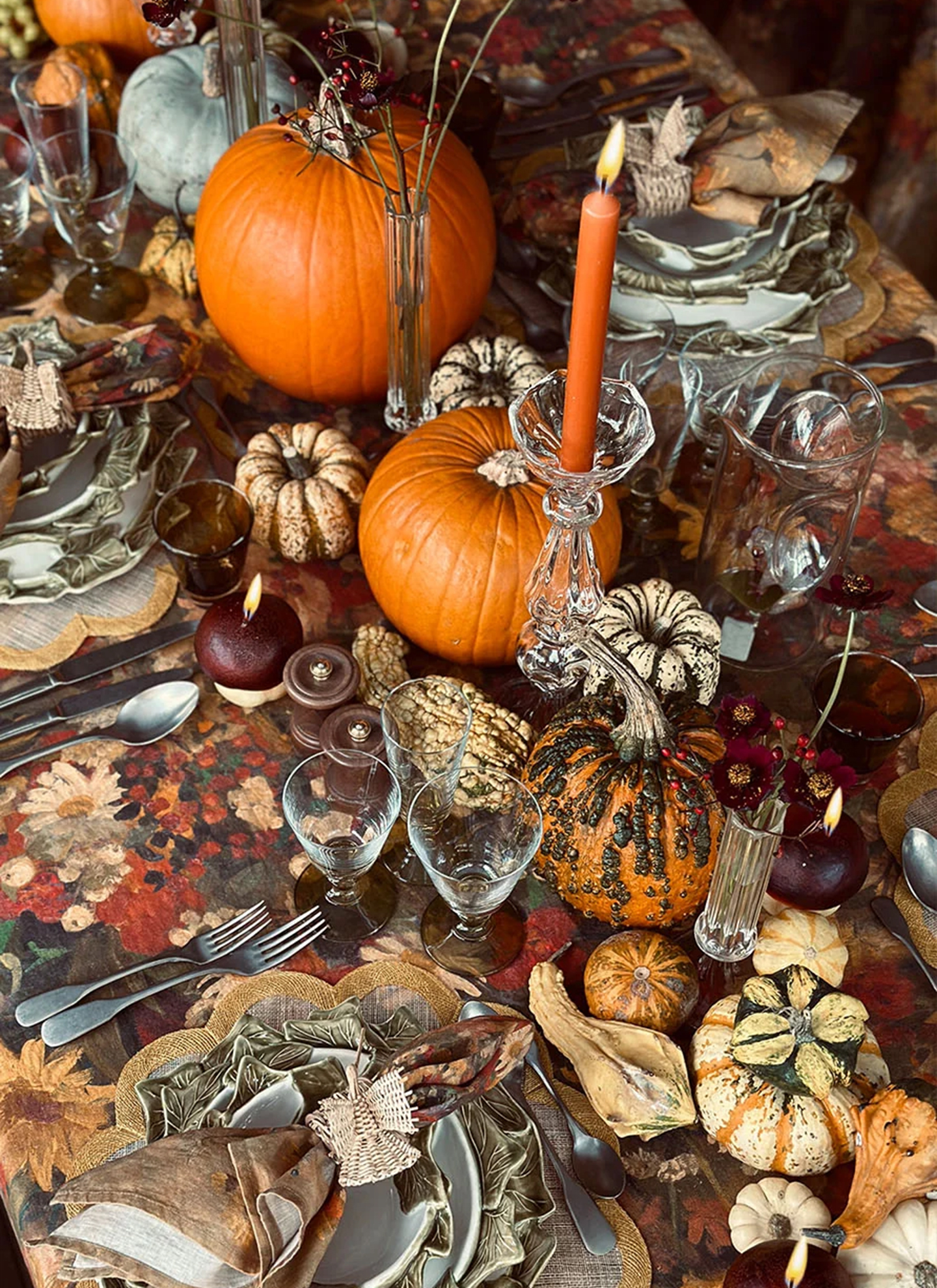 a close up of an autumn table with lots of real pumpkins, gourds and squashes, a dark floral tablecloth and natural textures and glassware