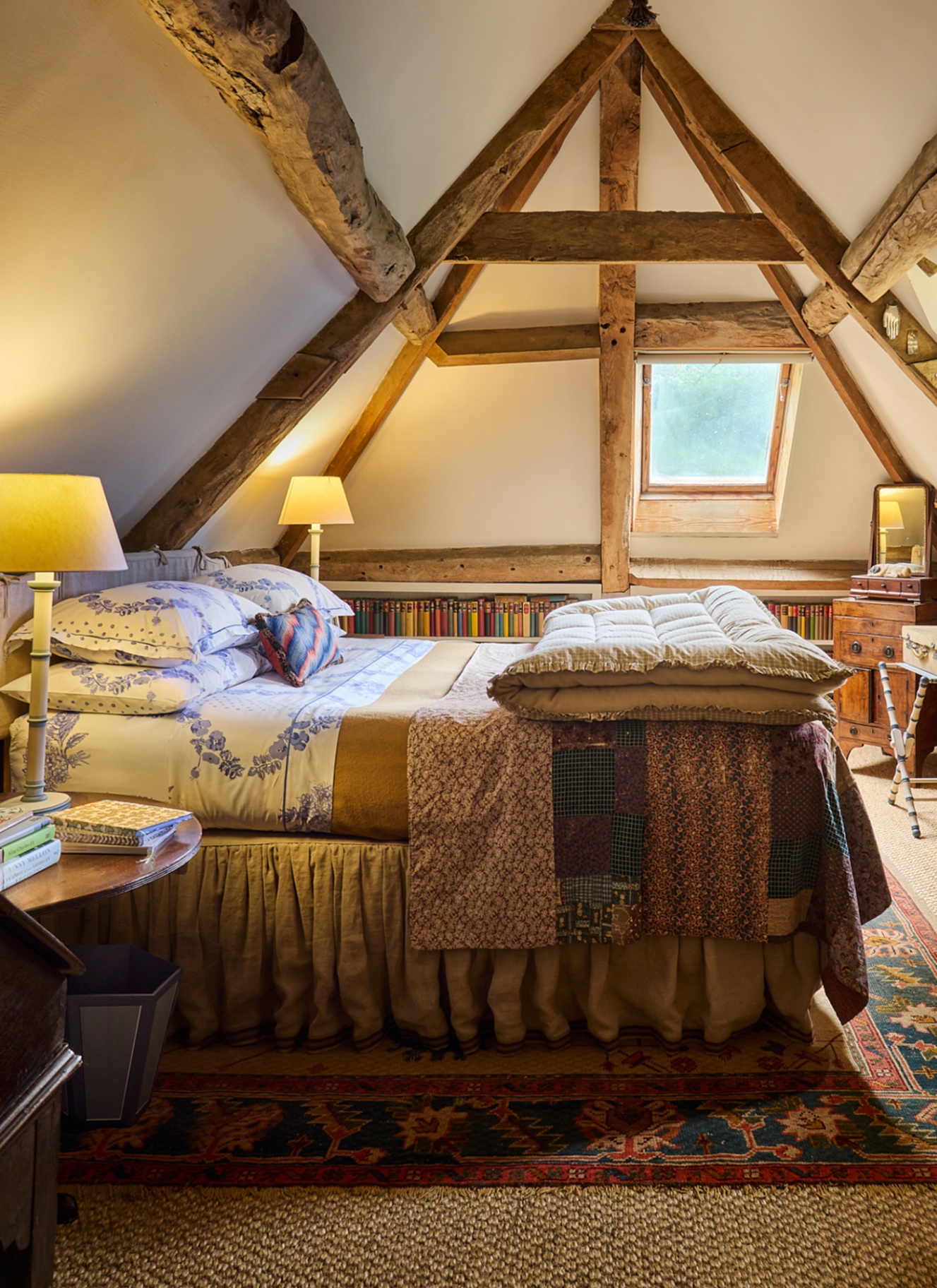 a bedroom with wooden beams on the ceiling, a large bed with white and blue floral bedding and a patchwork bedspread, low level bookcase shelves and white table lamps