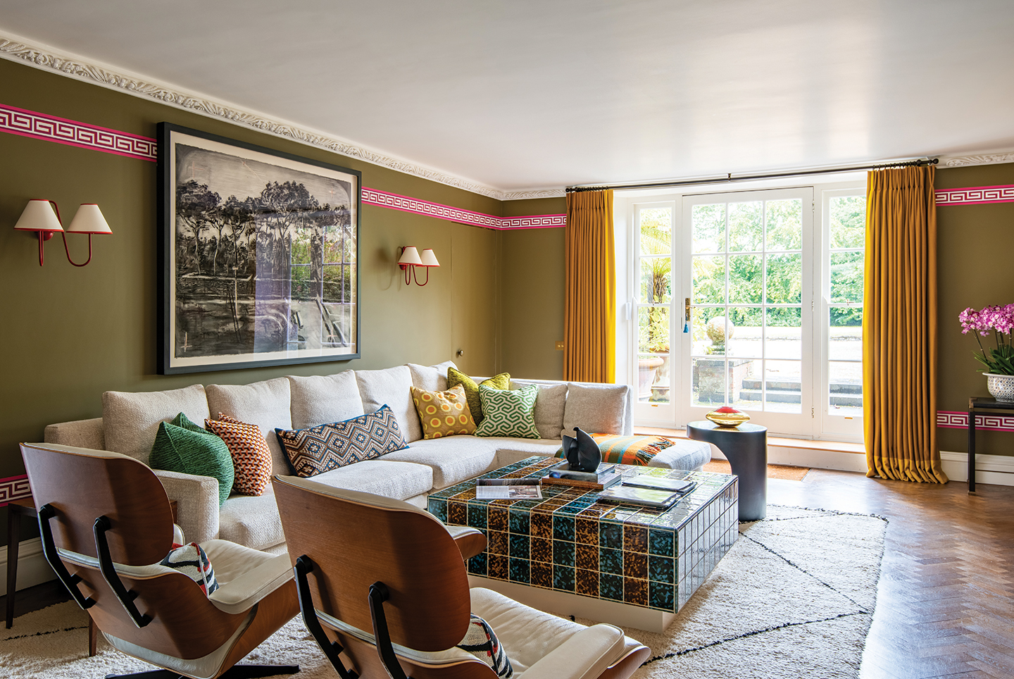 an informal living room with olive green walls with a greek key dado border and mustard yellow curtains. In the centre of the room is an L-shape white sofa dressed with patterned cushions, two mid-century chairs and a tiled coffee table