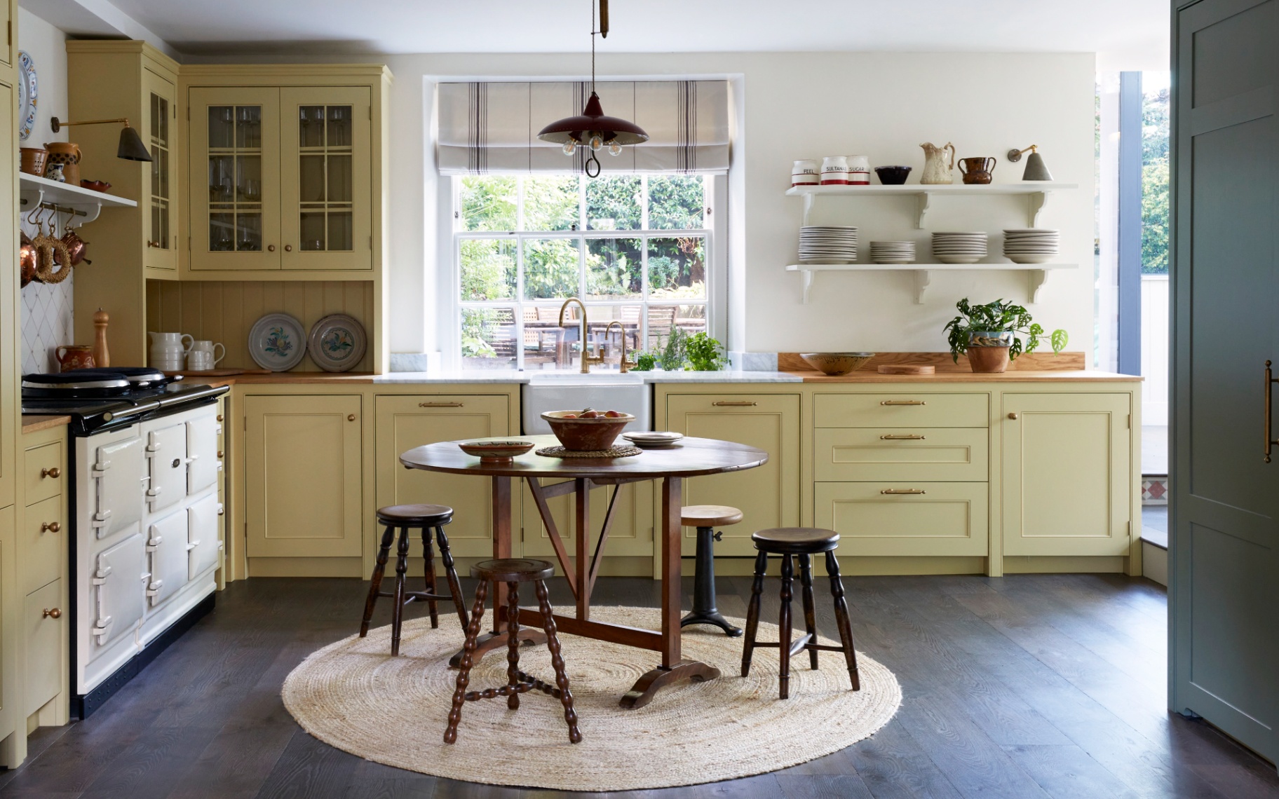 a kitchen with pale yellow cabinetry, a white range cooker and a round dining table with stools on a round jute rug