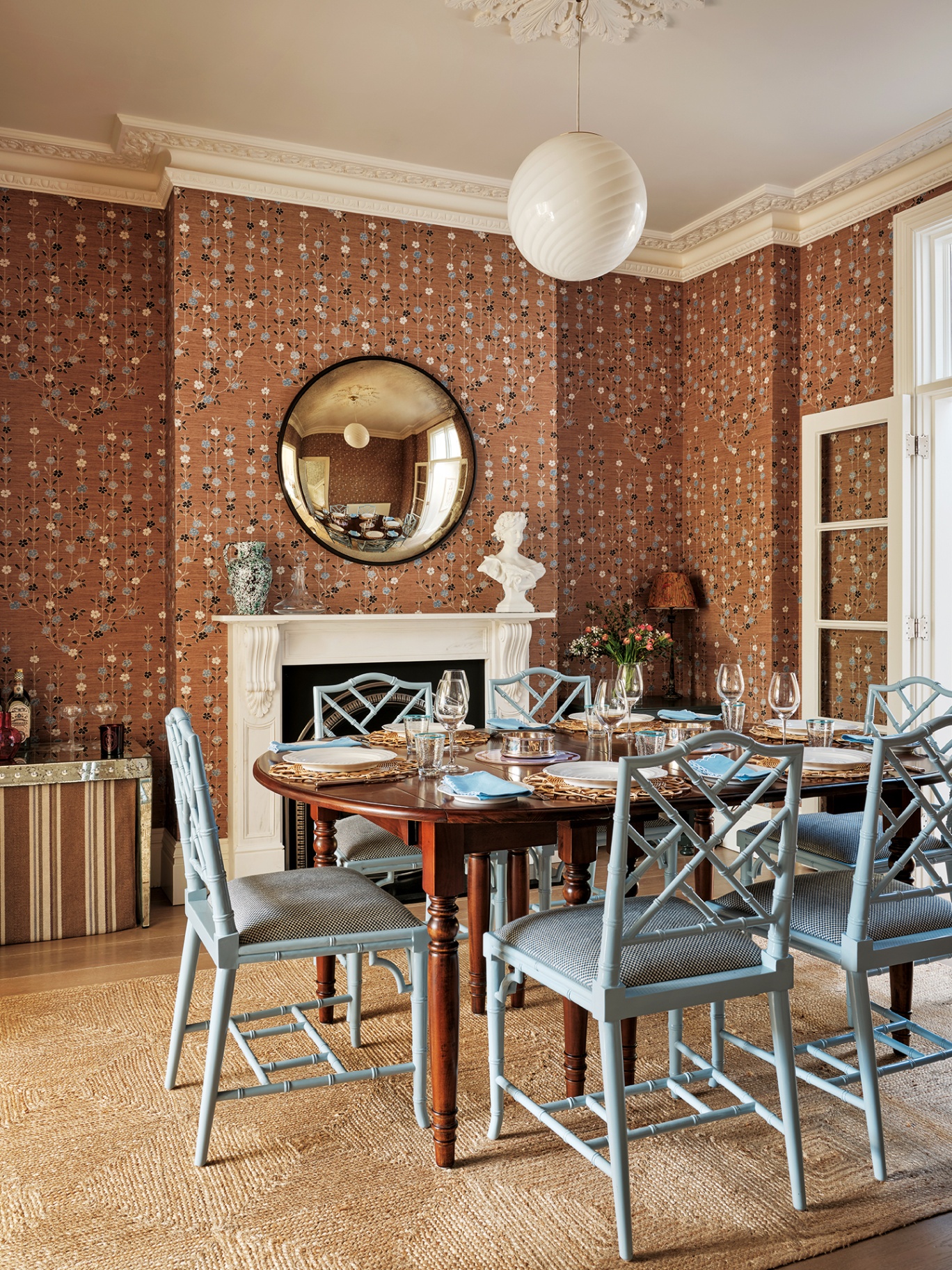 a dining room with brown floral wallpaper, and a wooden table in the centre surrounded by blue faux bamboo lacquer dining chairs. On the back wall is an ornate fireplace and above hangs a circular convex mirror