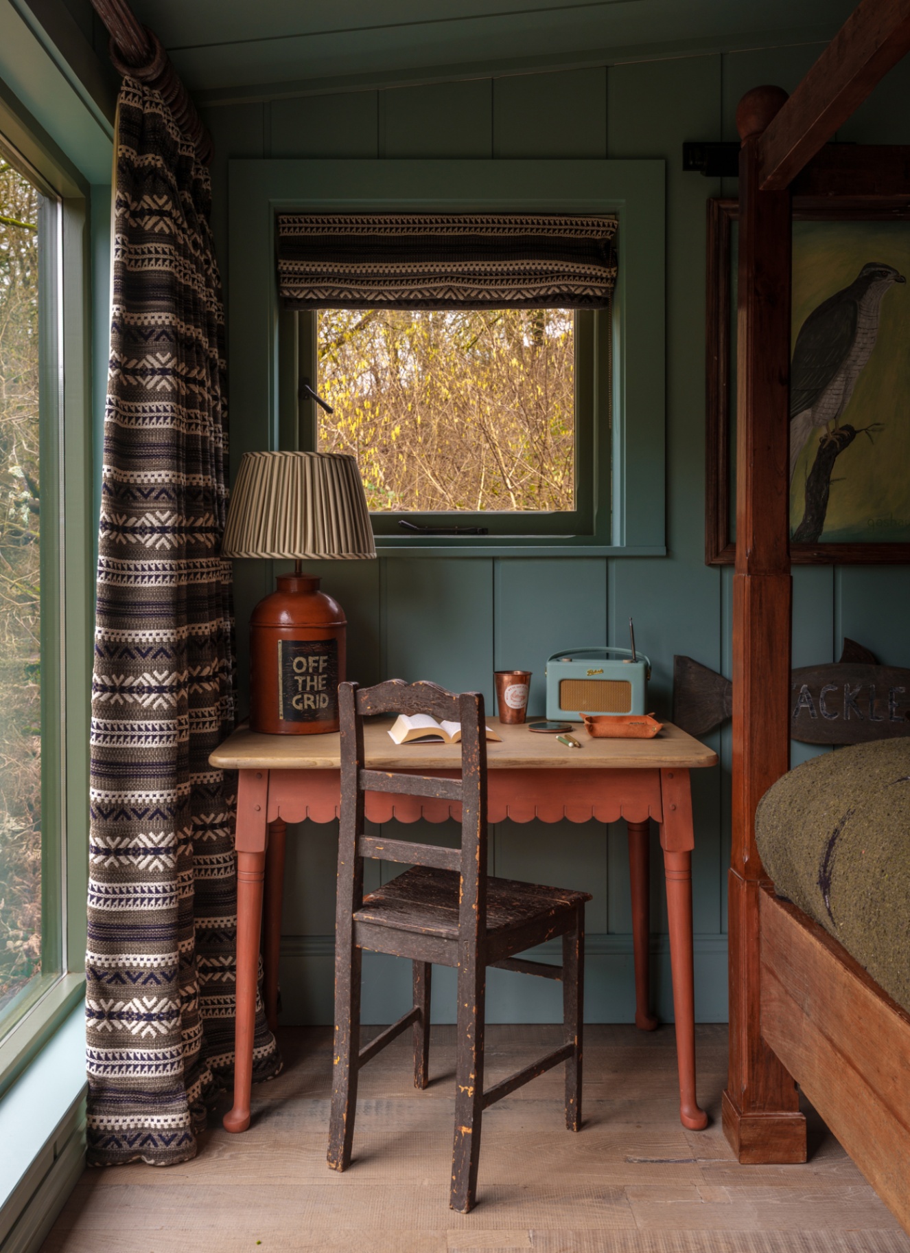 Cozy rustic study corner with dark sage wood paneled walls, a small red desk, dark wooden chair, table lamp with a striped shade, and patterned window drapes