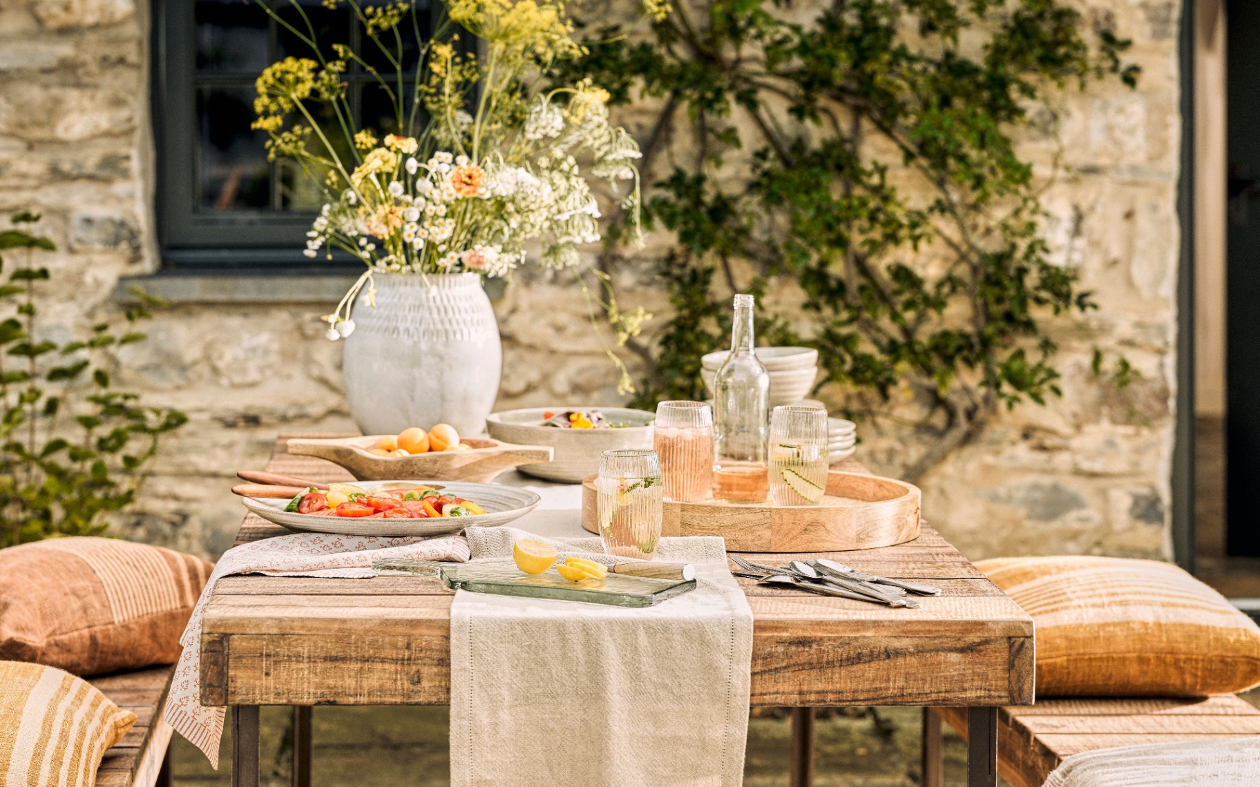 an outdoor wooden table with benches with orange cushions, cutlery and plates full of food and a large white vase full of flowers