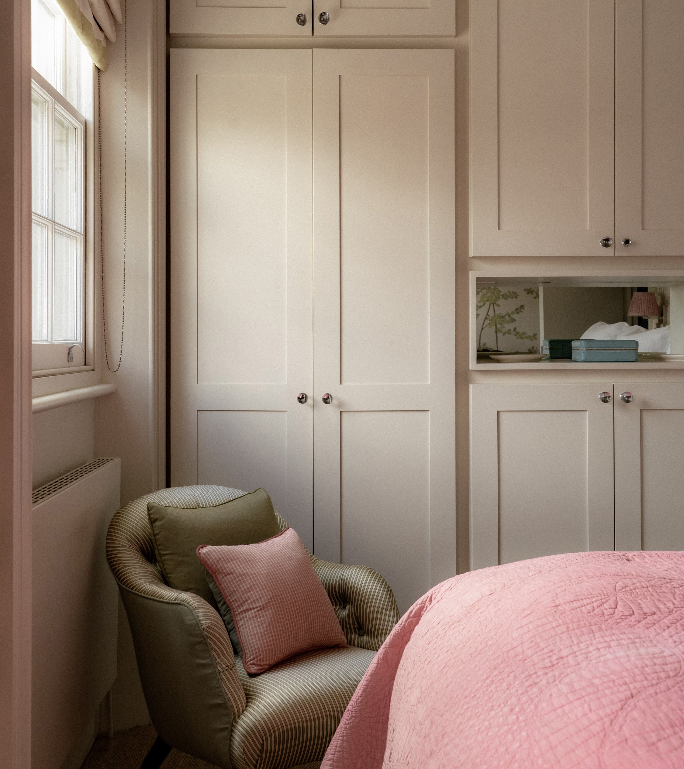 a bedroom with build in wardrobe painted in a neutral cream with a beige armchair in front with a pink cushion