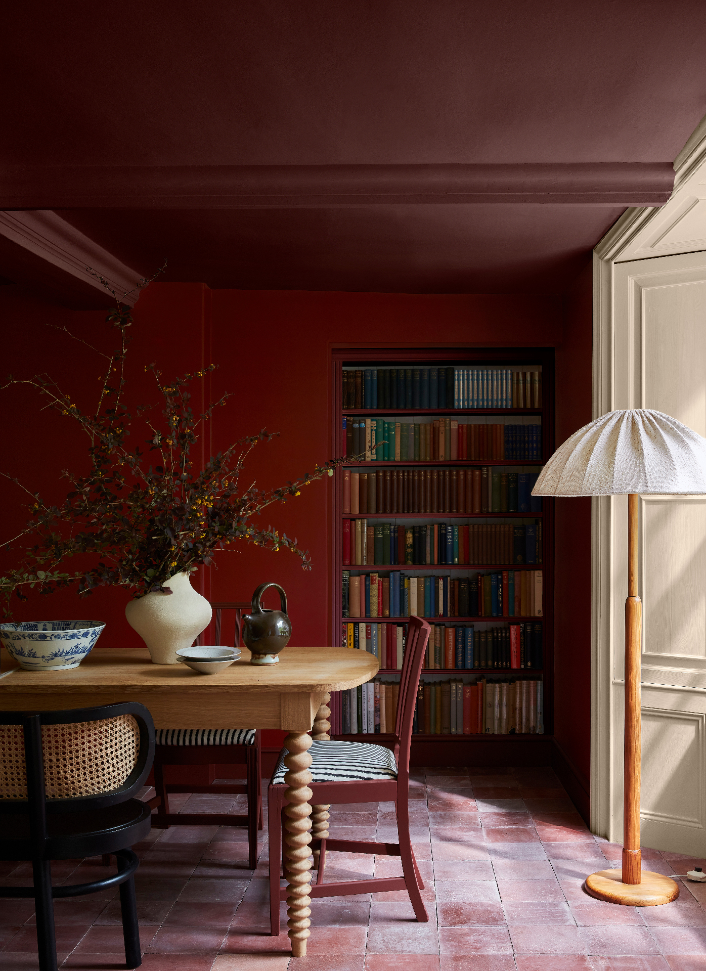 a dining room with dark red colour drenched walls and ceiling, a wooden dining table with bobbin legs and red chairs and a tall red bookcase