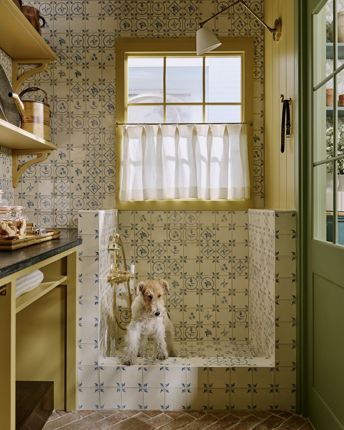 a utility room with a delft tiled dog wash station and a wire haired terrier inside, with yellow painted window and cabinetry