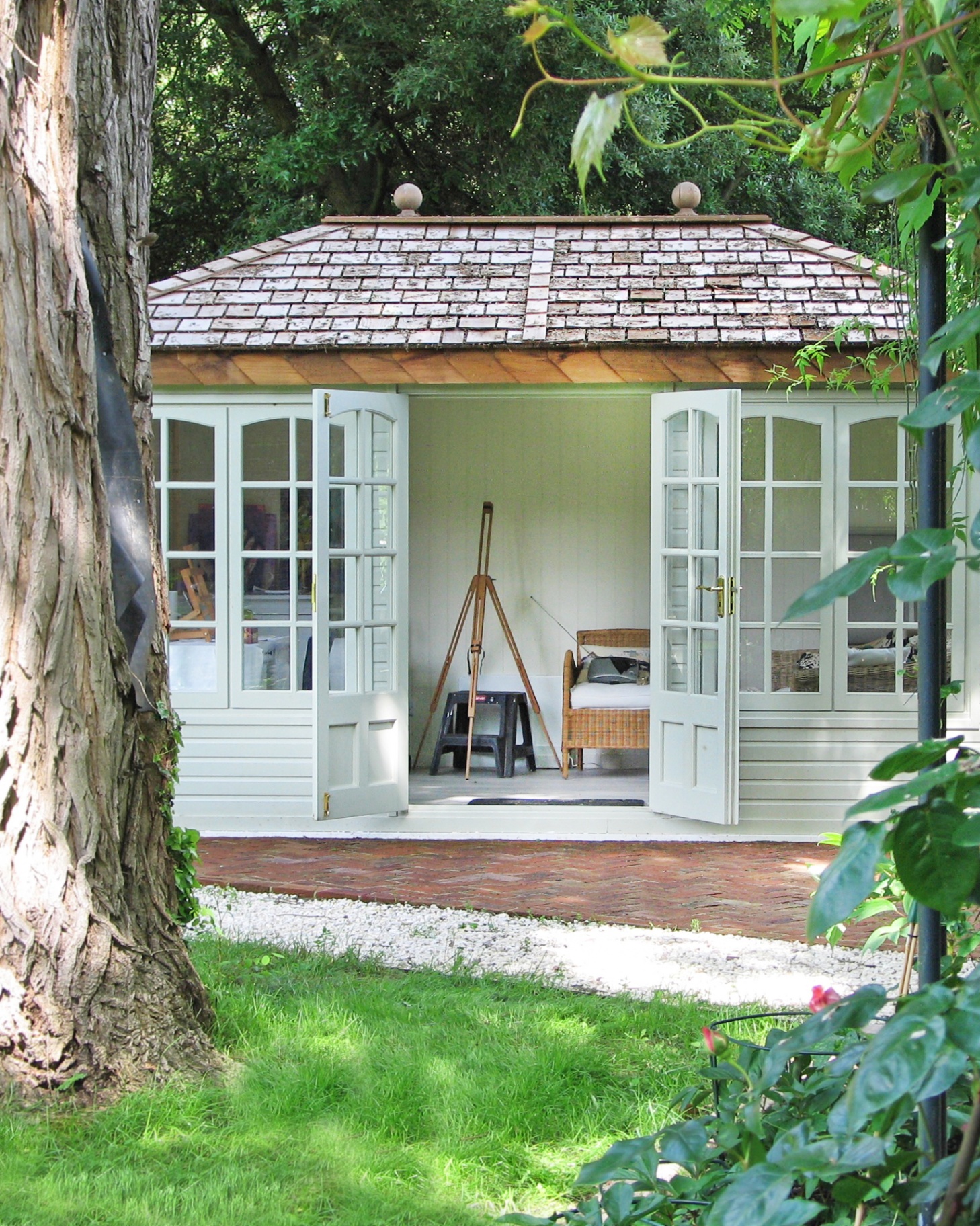 a sage green garden room with atiled roof with open doors looking into the room with an artists easel and a rattan sofa