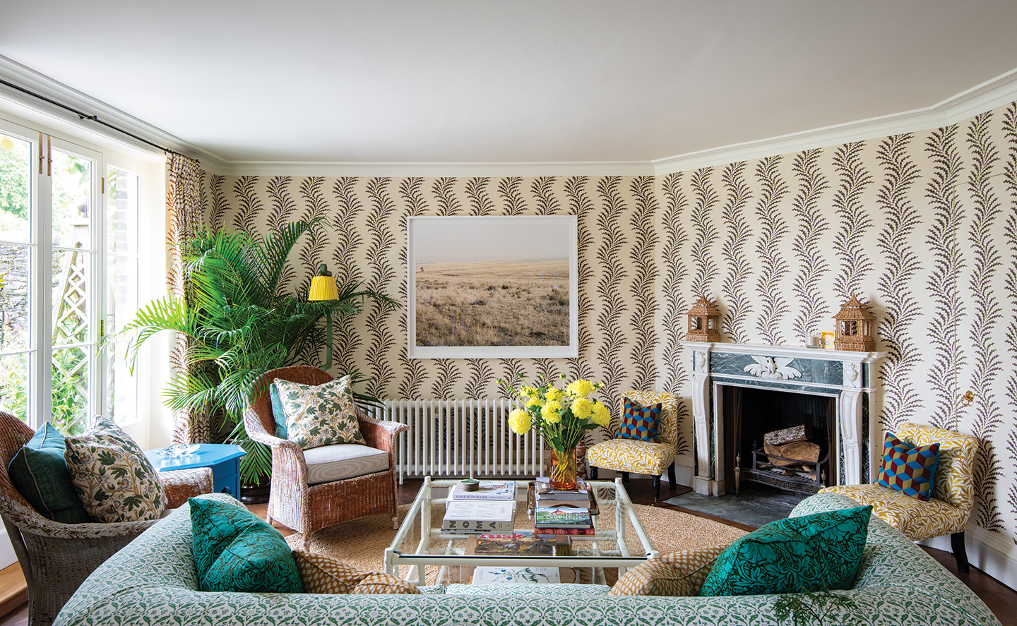 a garden room with sofa, armchairs and glass coffee table, with walls covered in a trailing vine wallpaper and an ornate blue stone fireplace to the right of the room. On the left are French windows and a large plant