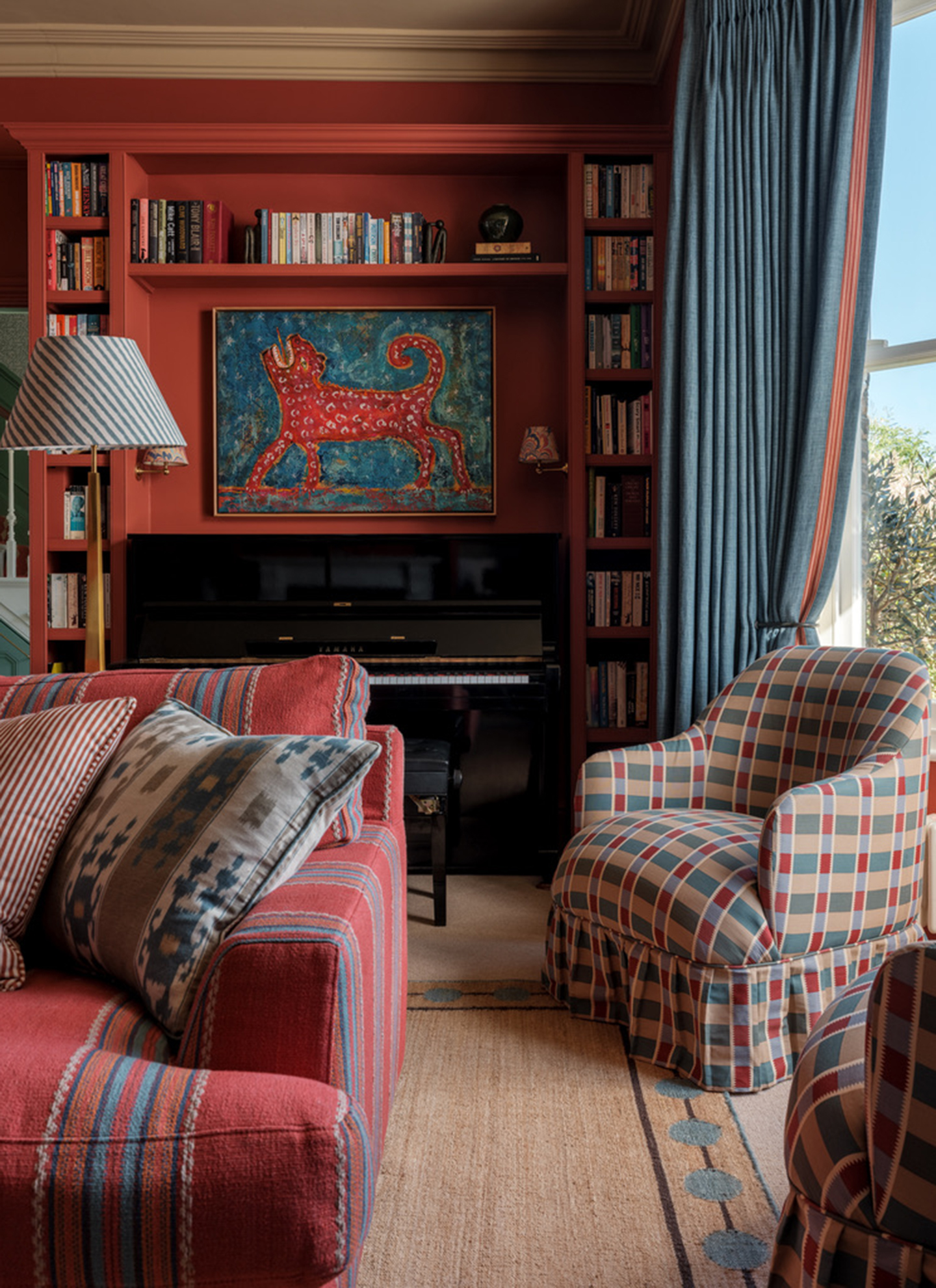 A warmly decorated living room featuring terracotta red built-in bookshelves, an upright piano, a red striped sofa, a plaid slipper chair, and blue curtains beside a window