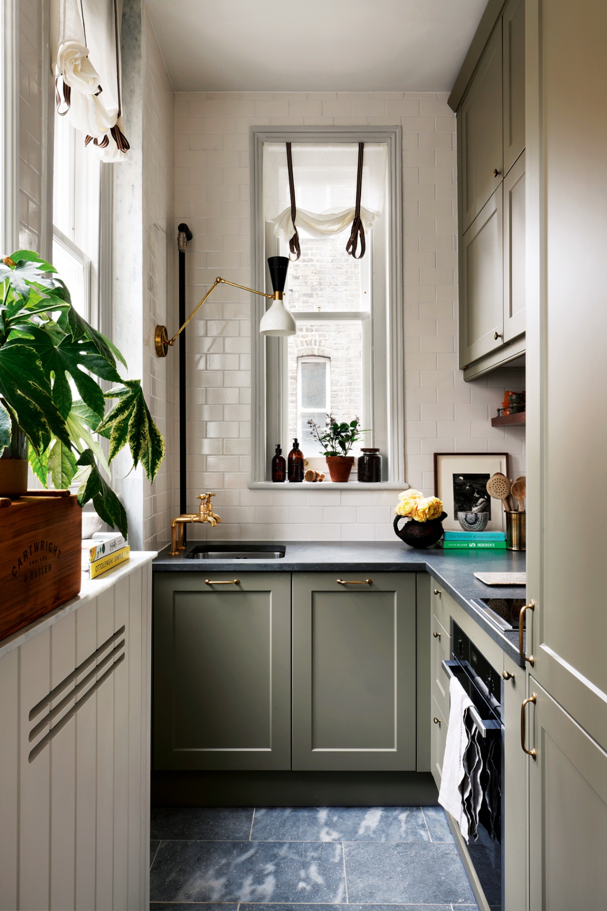 A tiny kitchen with grey cabinets finished with brass hardware, narrow windows in the background and to the left dressed with sheer blinds, a large wall light and a small sink paired with brass taps. The worktops are decorated with flowers and utensils