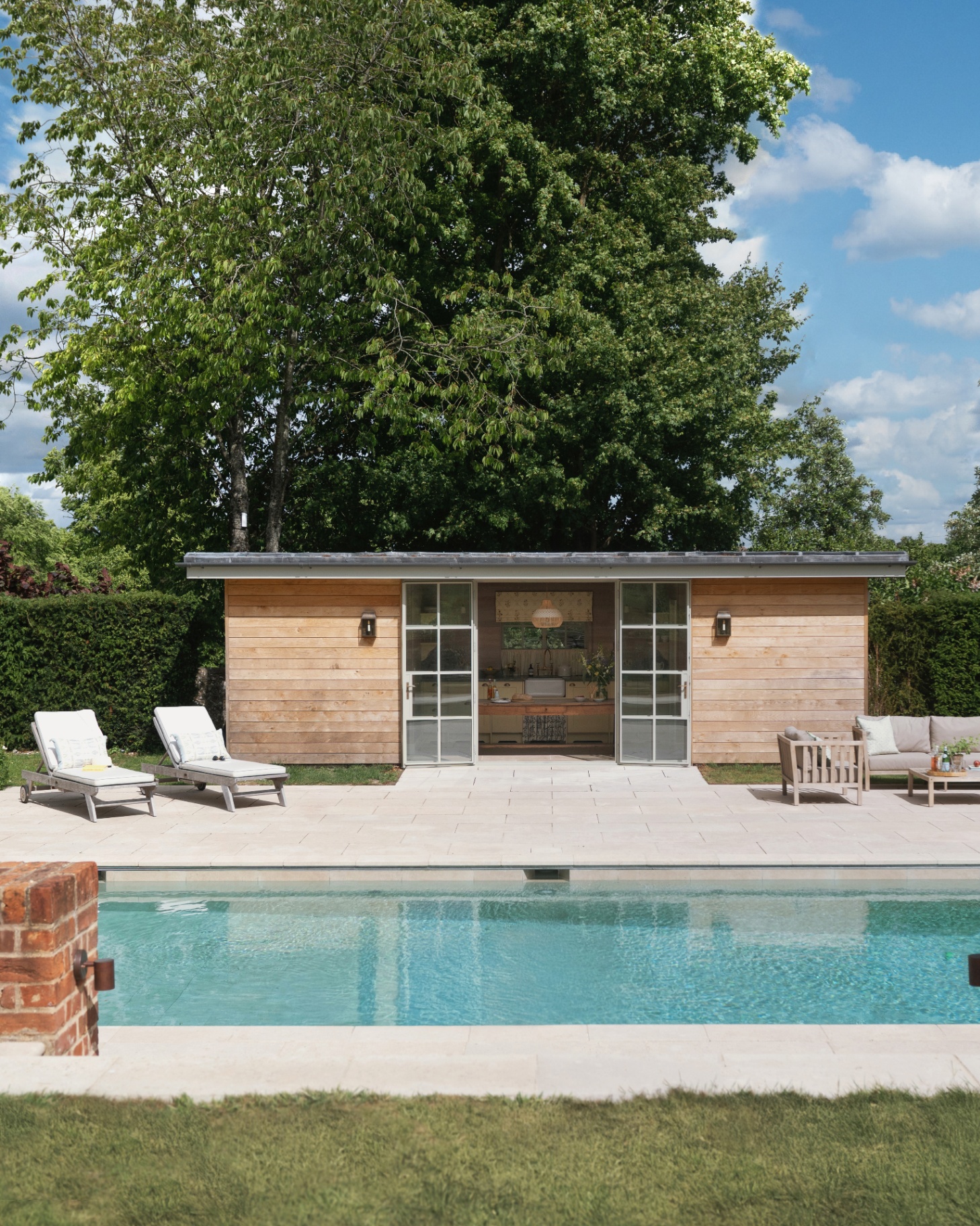 a wooden pool house with a pale grey patio and two sun loungers looking out to a large swimming pool