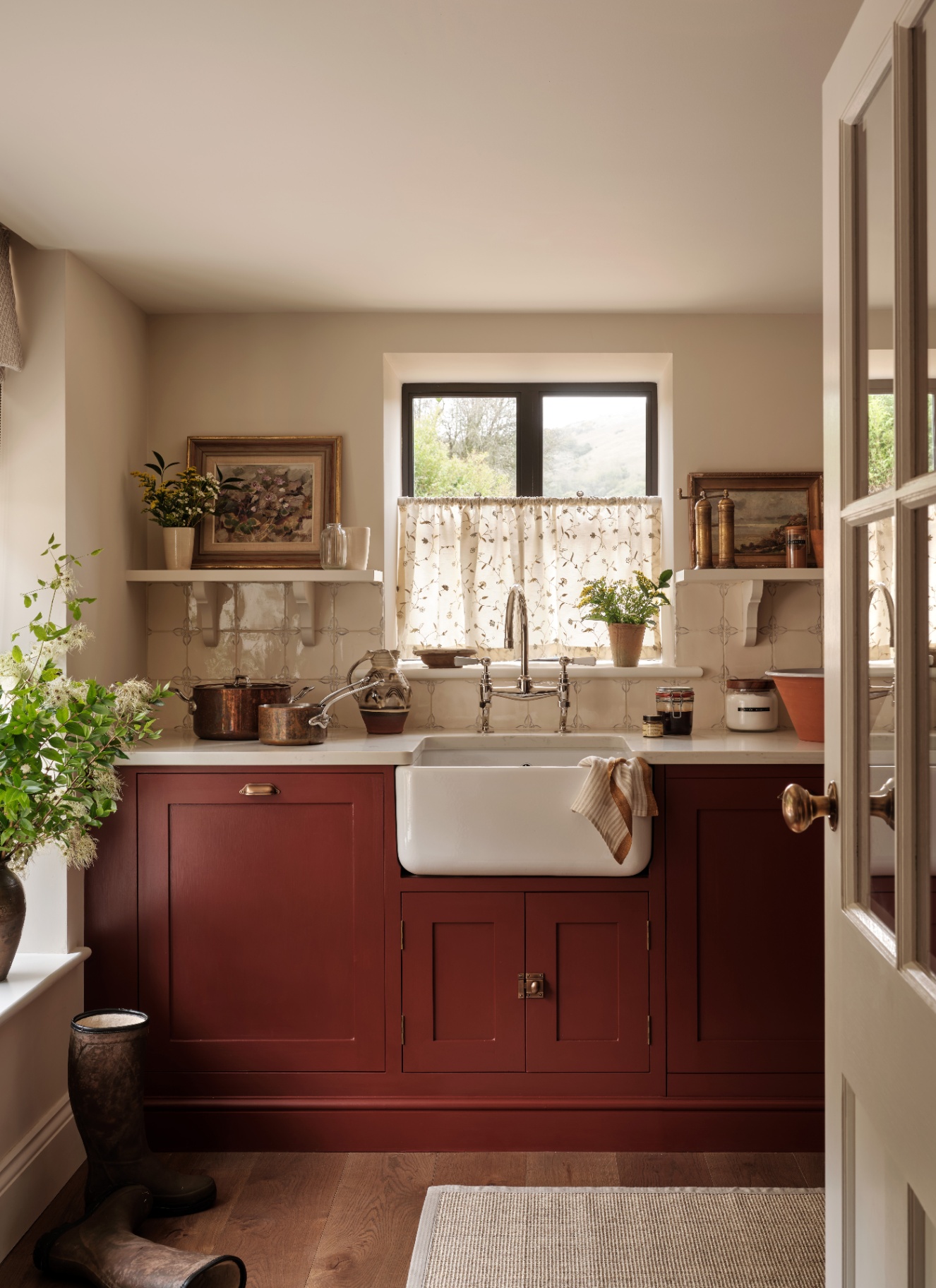 a country kitchen with deep red cabinetry, two shelves eitherside of the window with a gold framed painting and a cafe curtain on the window