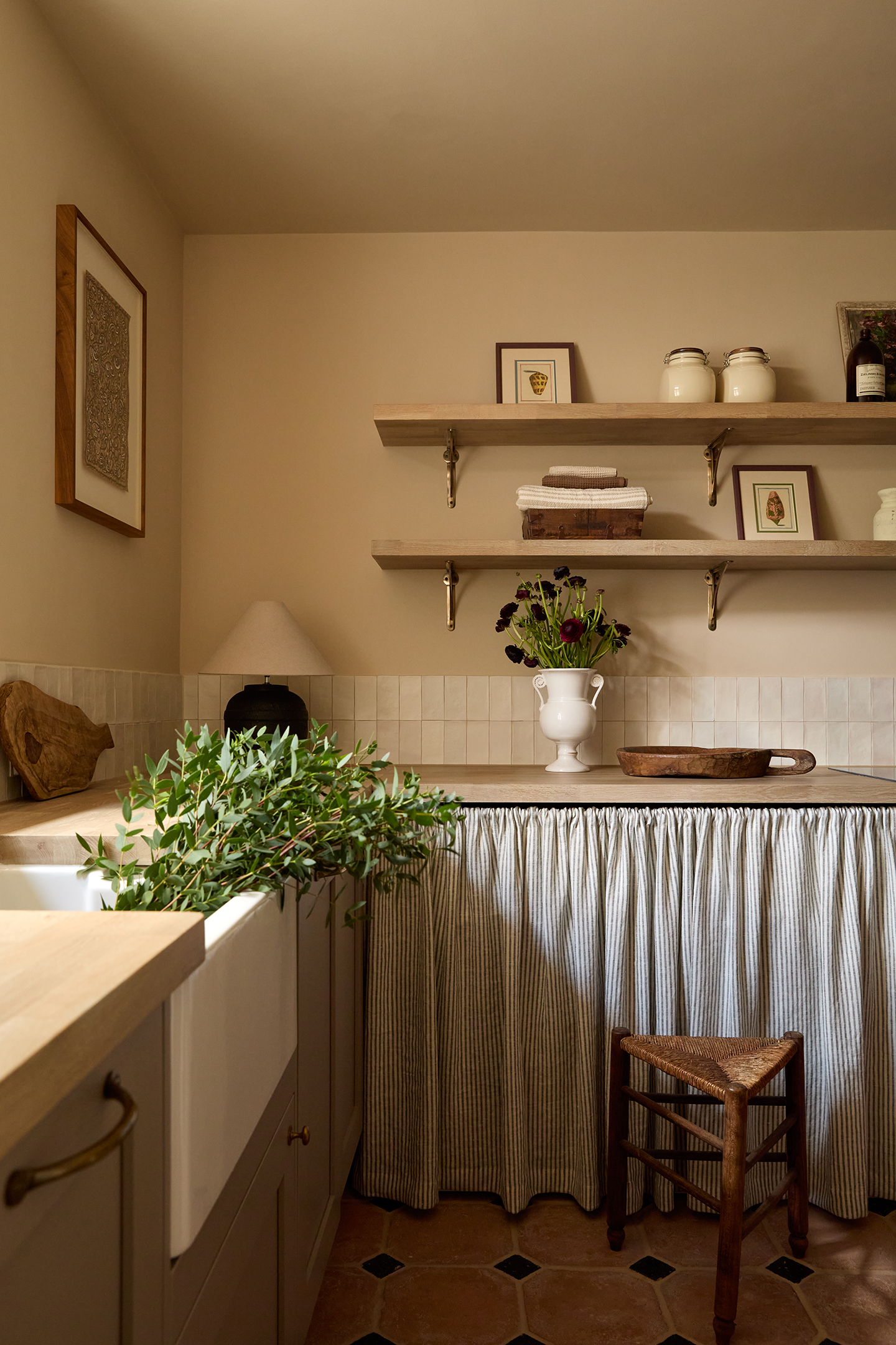 a utility room with neutral walls and a short white tiled backsplash. A sink to the left is filled with greenery. A sink skirt in ticking fabric runs along the back cabinets. Above open shelving is styled with artworks, kitchen textiles and ceramics